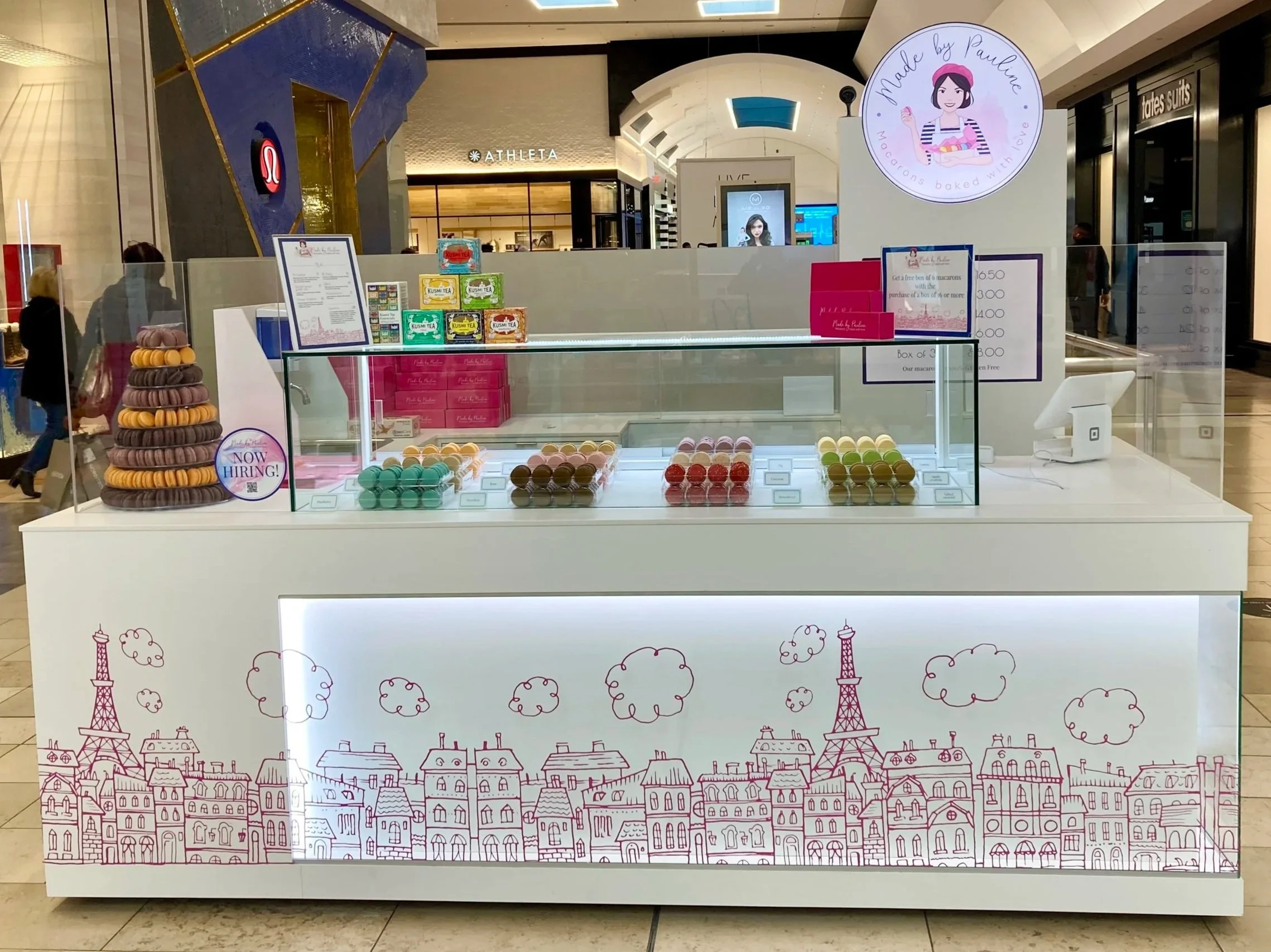A custom made macaron kiosk Made By Pauline Macarons with colorful macarons in front of a Paris-themed backdrop. The backdrop features illustrations of Parisian buildings and the Eiffel Tower with pink accents. located in Westfield Garden State Plaza