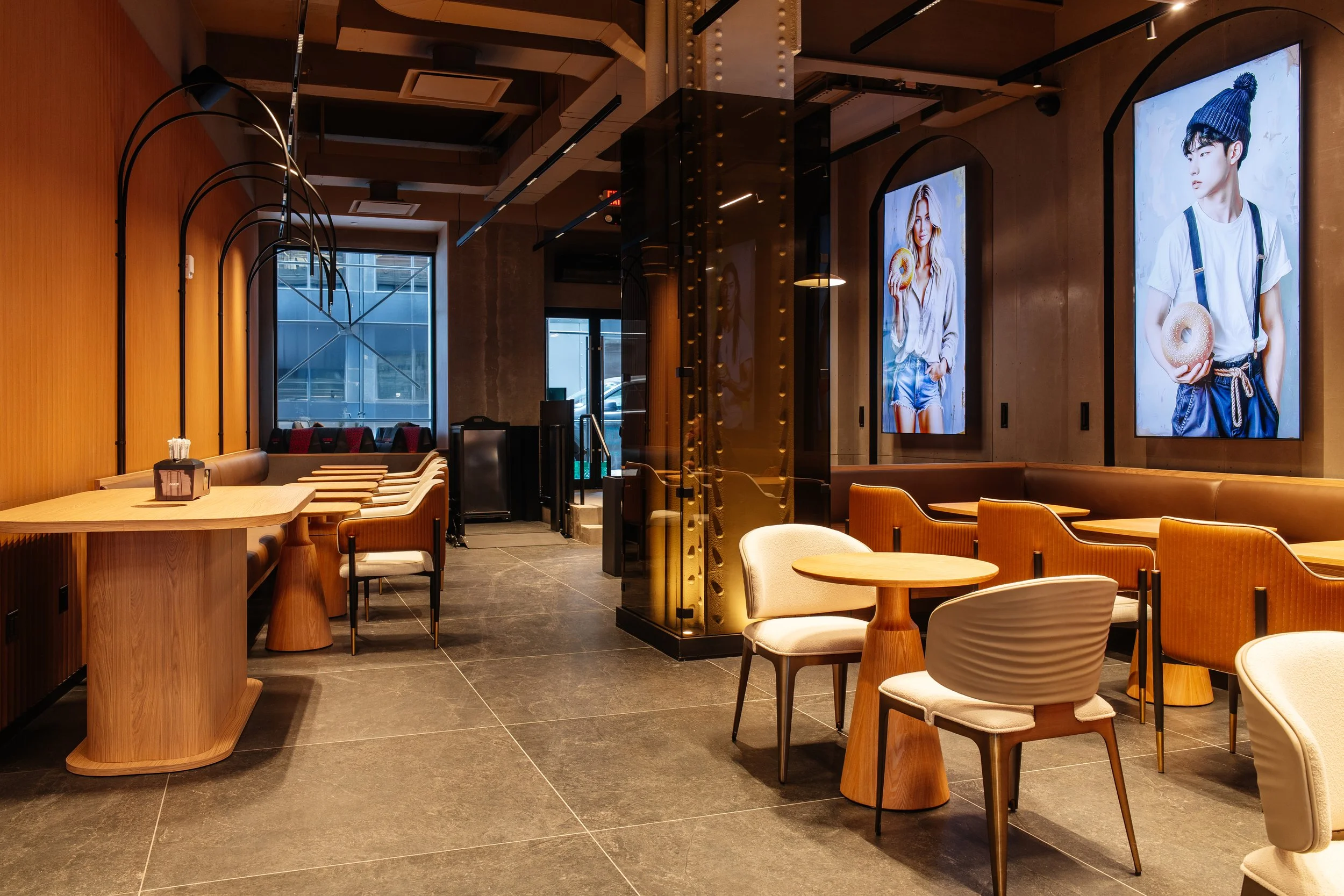 Boobliq Bagel Bistro Interior of a modern restaurant or cafe with wooden decor, stylish chairs and tables, large digital screens displaying fashion models, and large windows providing natural light.