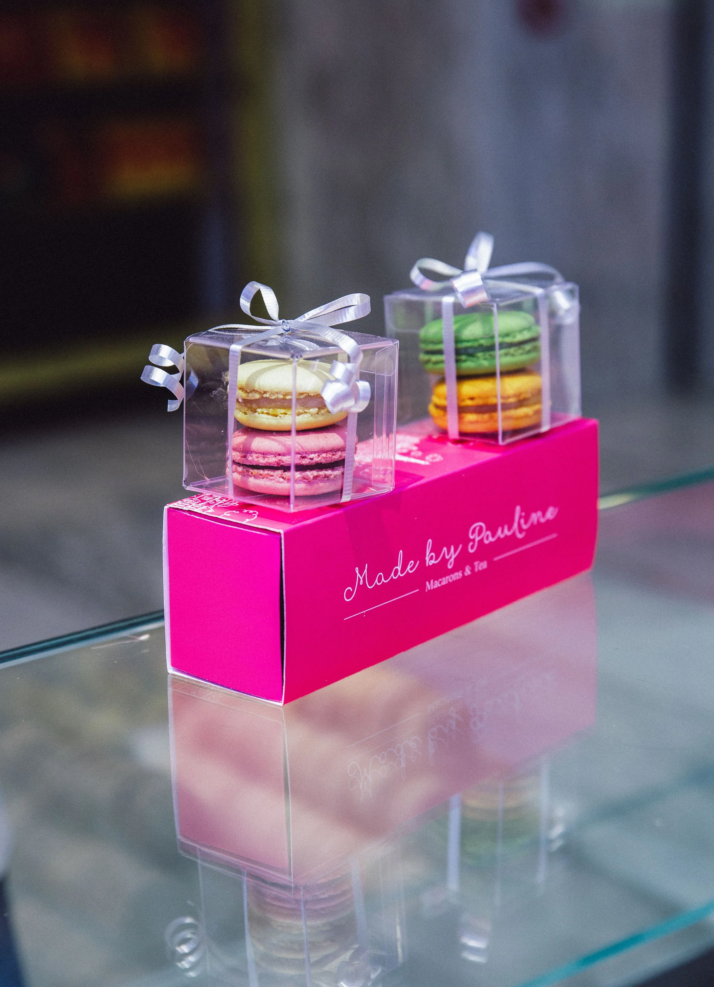 Colorful macarons in clear plastic gift boxes with white ribbons, placed on a pink "Made by Pauline" box.