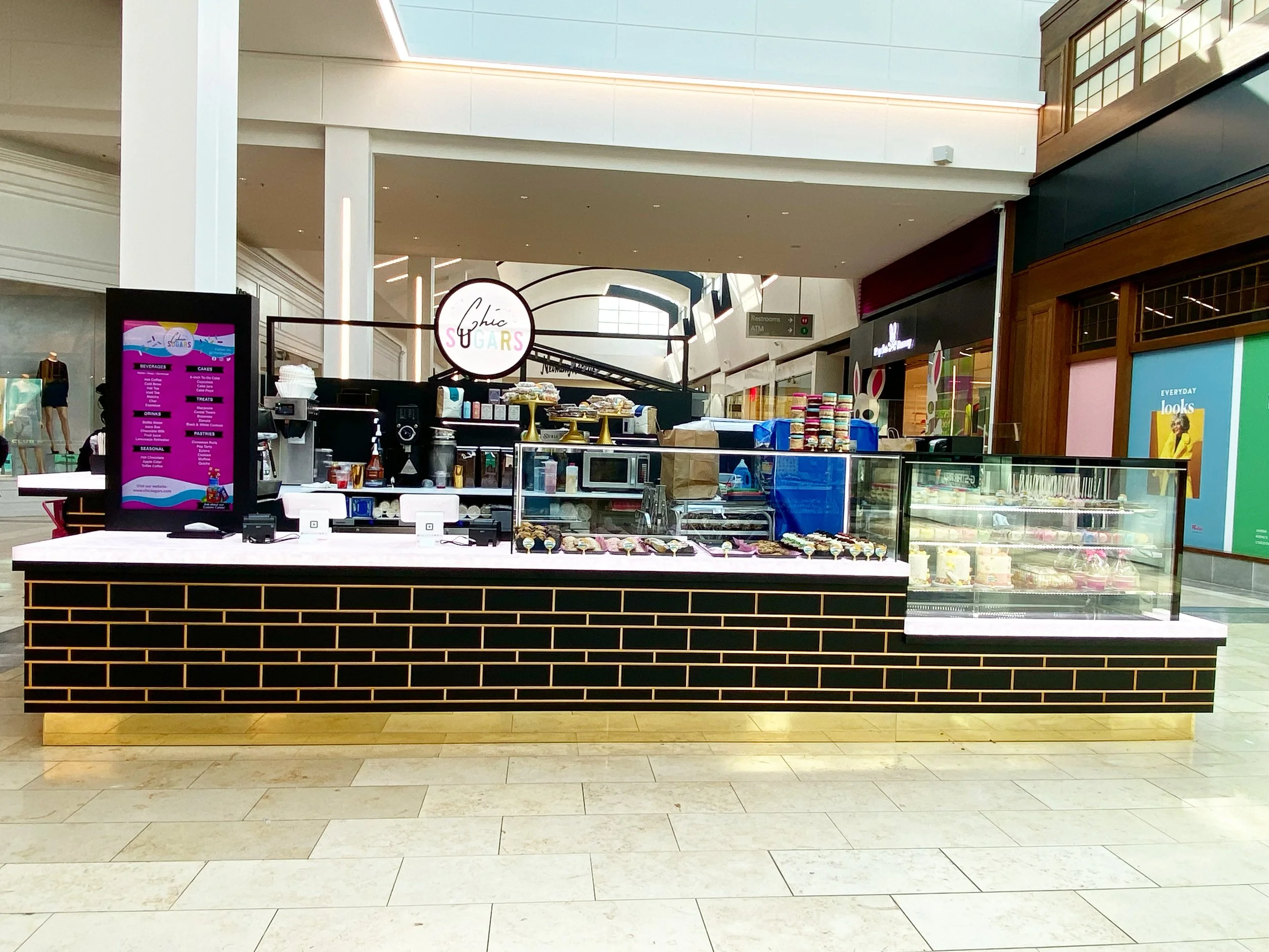 A kiosk inside a shopping mall Westfield Garden State Plaza, NJ with a sign reading 'Chic Sugars' cake studio, displaying baked goods and desserts, with a digital menu board on the left.