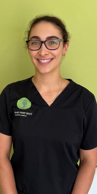 Smiling woman with glasses wearing a black shirt with a logo that says 'The Foot Spot' against a light green background.