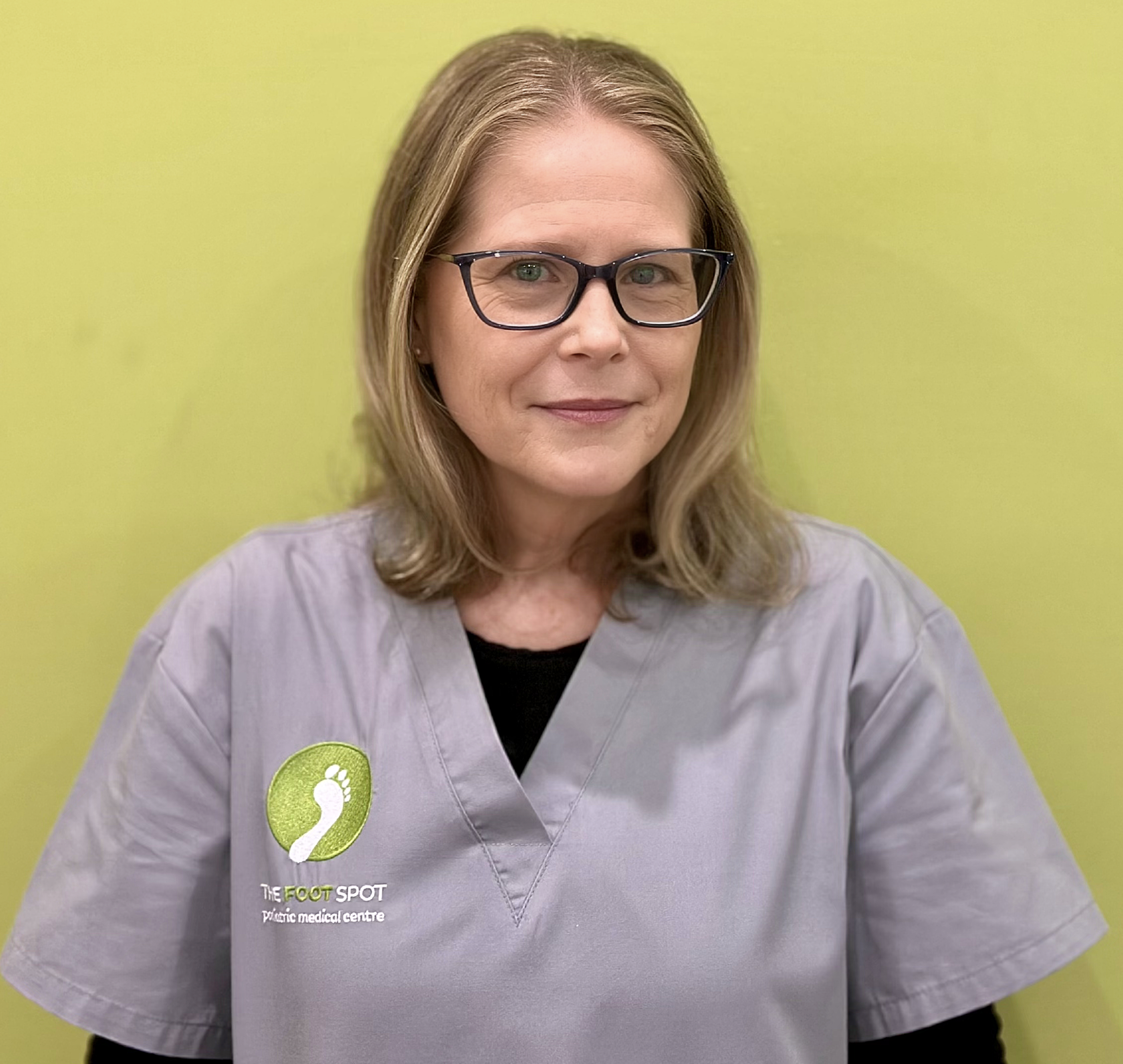 A woman with shoulder-length blonde hair, glasses, and wearing a gray medical uniform with a logo that says 'The Foot Spot Piaggio Medical Centre' standing in front of a yellow-green wall.
