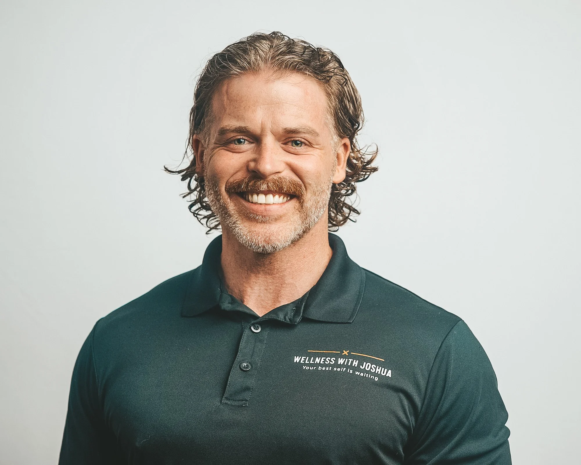 A smiling middle-aged man with wavy, grayish hair and a beard, wearing a black polo shirt with the text 'WELLNESS WITH JOSHUA Your best self is waiting' on it, standing against a plain light gray background.