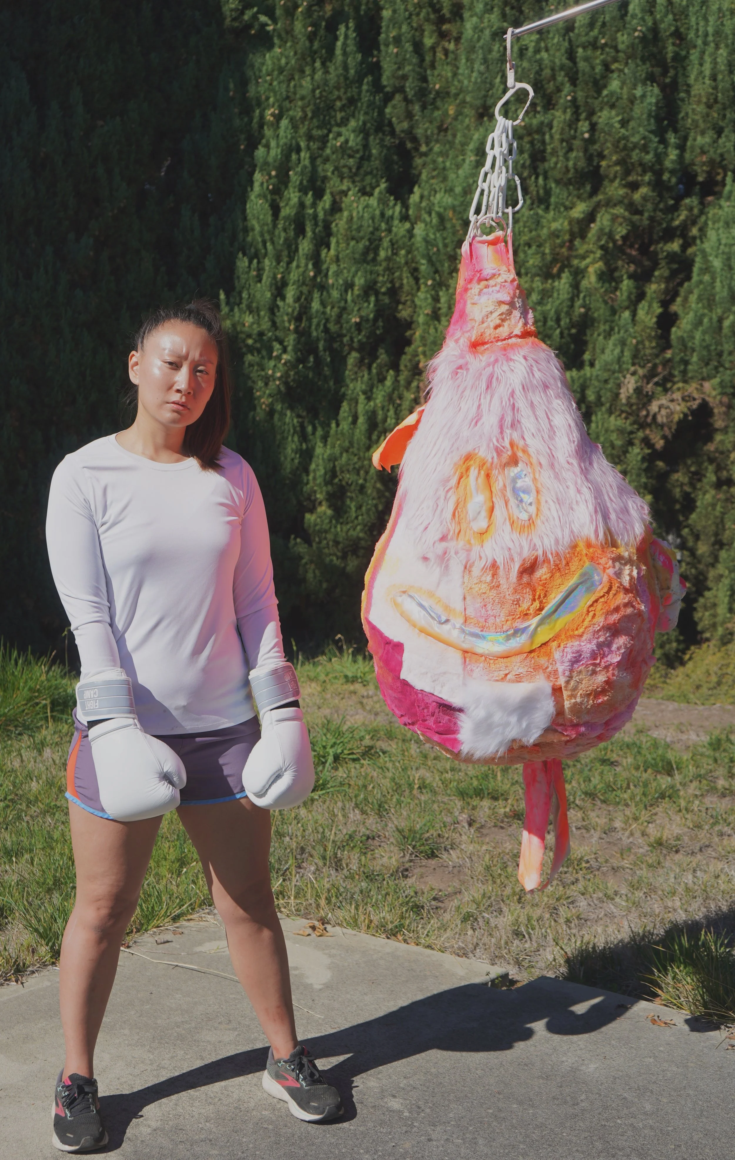 The Recovering People Pleaser sculpture by Sophia Huang, hanging mixed media sculpture with plush and resin elements, shown outdoors beside the artist wearing boxing gloves