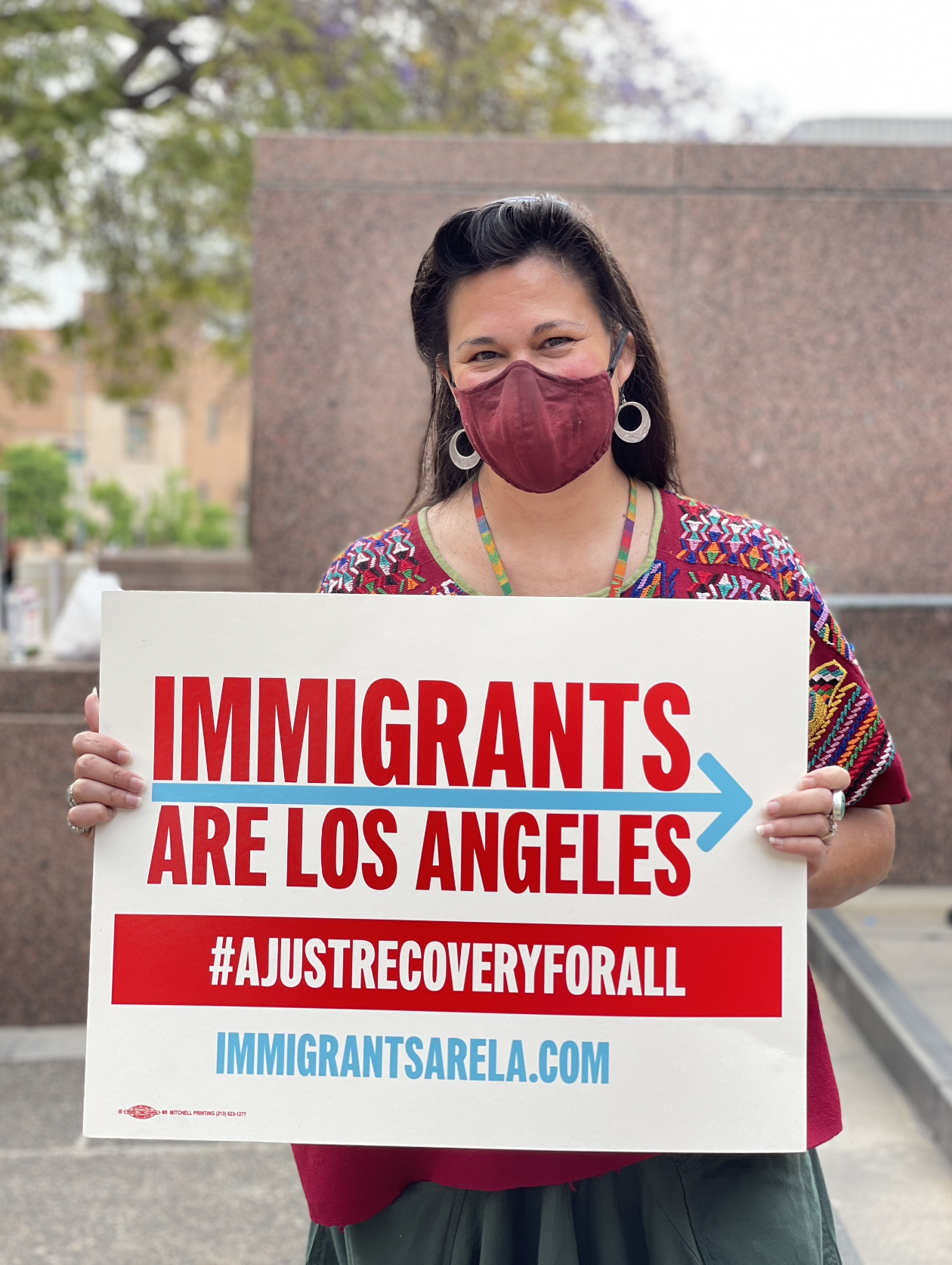 A woman wearing a maroon face mask and colorful embroidered top holding a sign that reads 'Immigrants are Los Angeles' with a hashtag #AjustRecoveryForAll and a website 'immigrantsarela.com'.