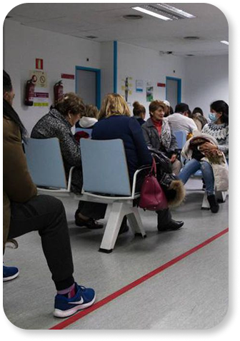 Varios adultos sentados en una sala de espera, algunos con mascarillas, frente a una pared blanca con carteles y un pasillo con línea roja en el suelo.