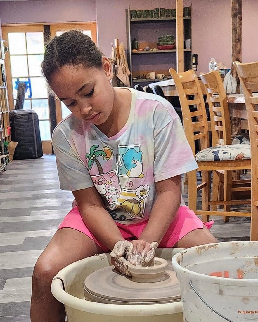 Five children standing in a pottery studio holding small pumpkin or Halloween-themed decorations. They are in front of metal shelves filled with various ceramic vases and pottery items.