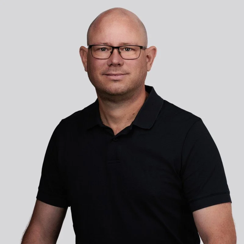 A man with glasses and a bald head wearing a black polo shirt against a plain gray background.