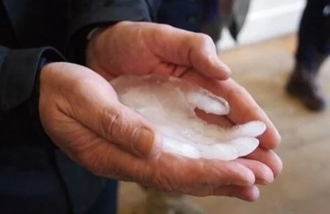 A man cups his hands to hold an ice cube in the shape of a hand  smaller than his with the palm facing upwards.