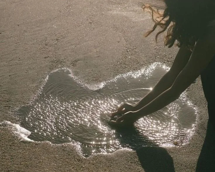 A woman squats down and cups her hand to scoop up water from a puddle at the beach.