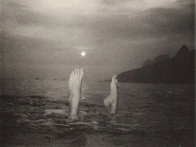 Two feet sticking out of the ocean's surface with the full moon pictured just above their right big toe.