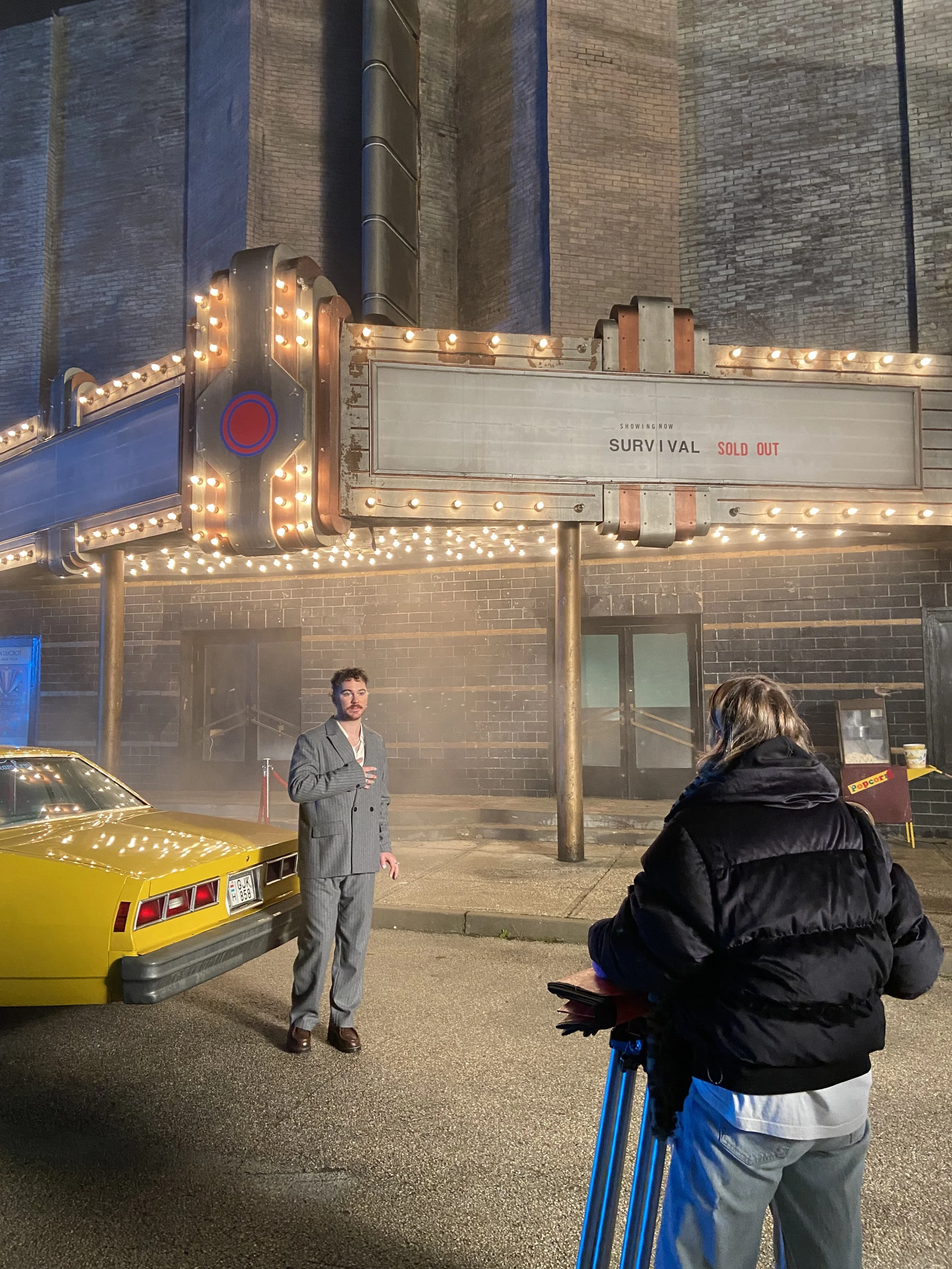 A film crew is shooting a scene outside a theater at night. One man in a gray suit is standing next to a yellow car, and a woman with a camera is filming him. The theater marquee shows "Survival," which is sold out, and is surrounded by bright marquee lights. The background includes a brick building and a popcorn stand.