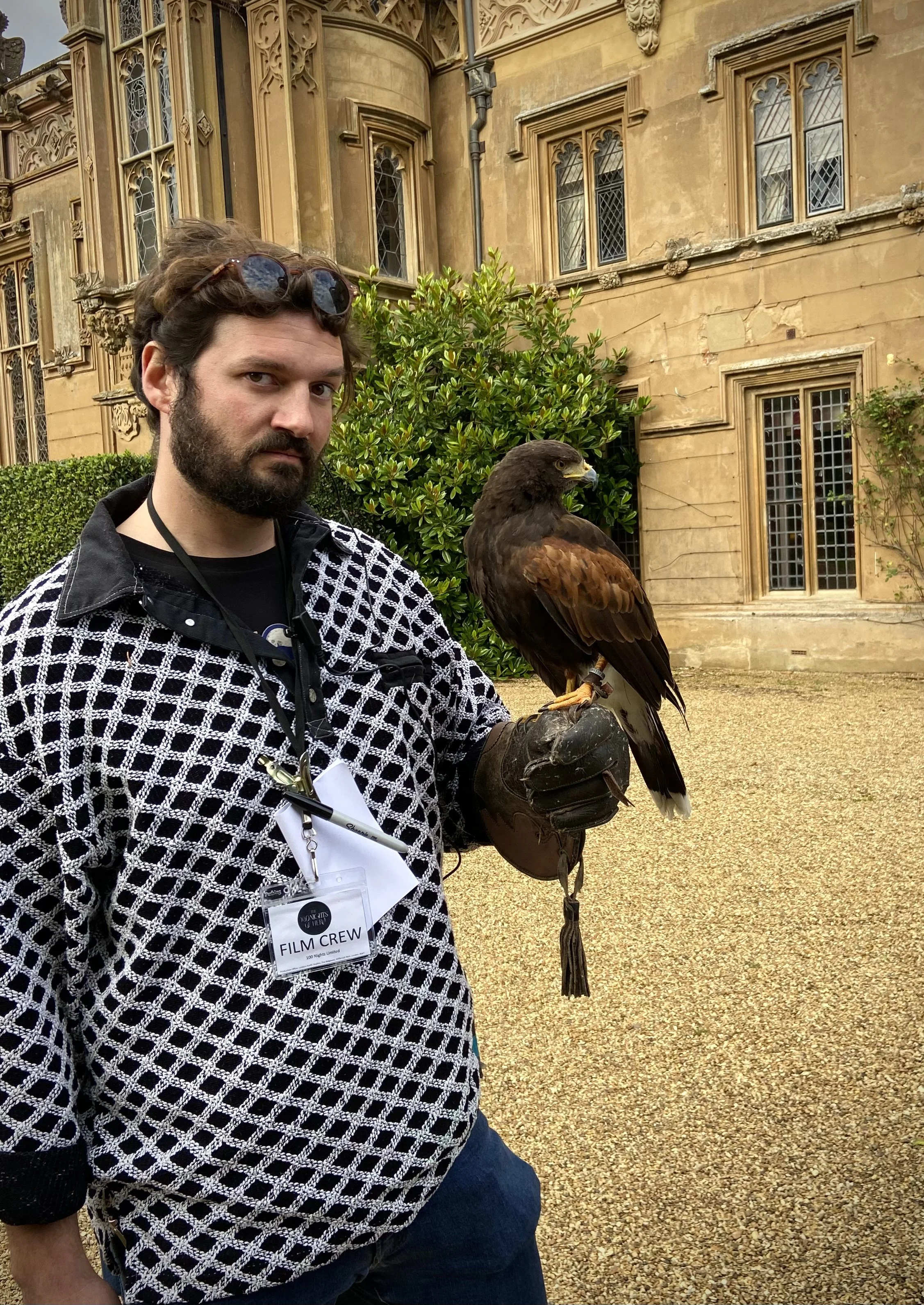 A man with sunglasses on his head, wearing a black and white patterned jacket, holds a bird of prey with a glove. The background shows an old, ornate building with large windows and green bushes.