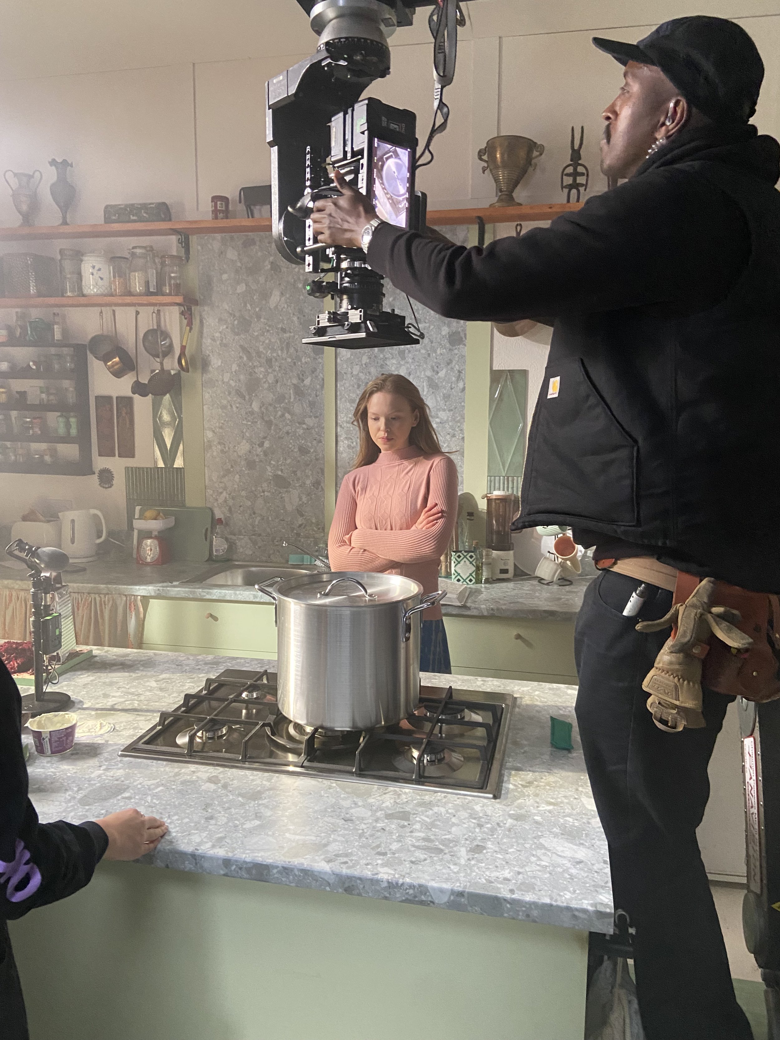 A woman in a pink sweater standing in a kitchen with her arms crossed, while a man operates filming equipment from above, capturing her on camera near a stove with a large pot.