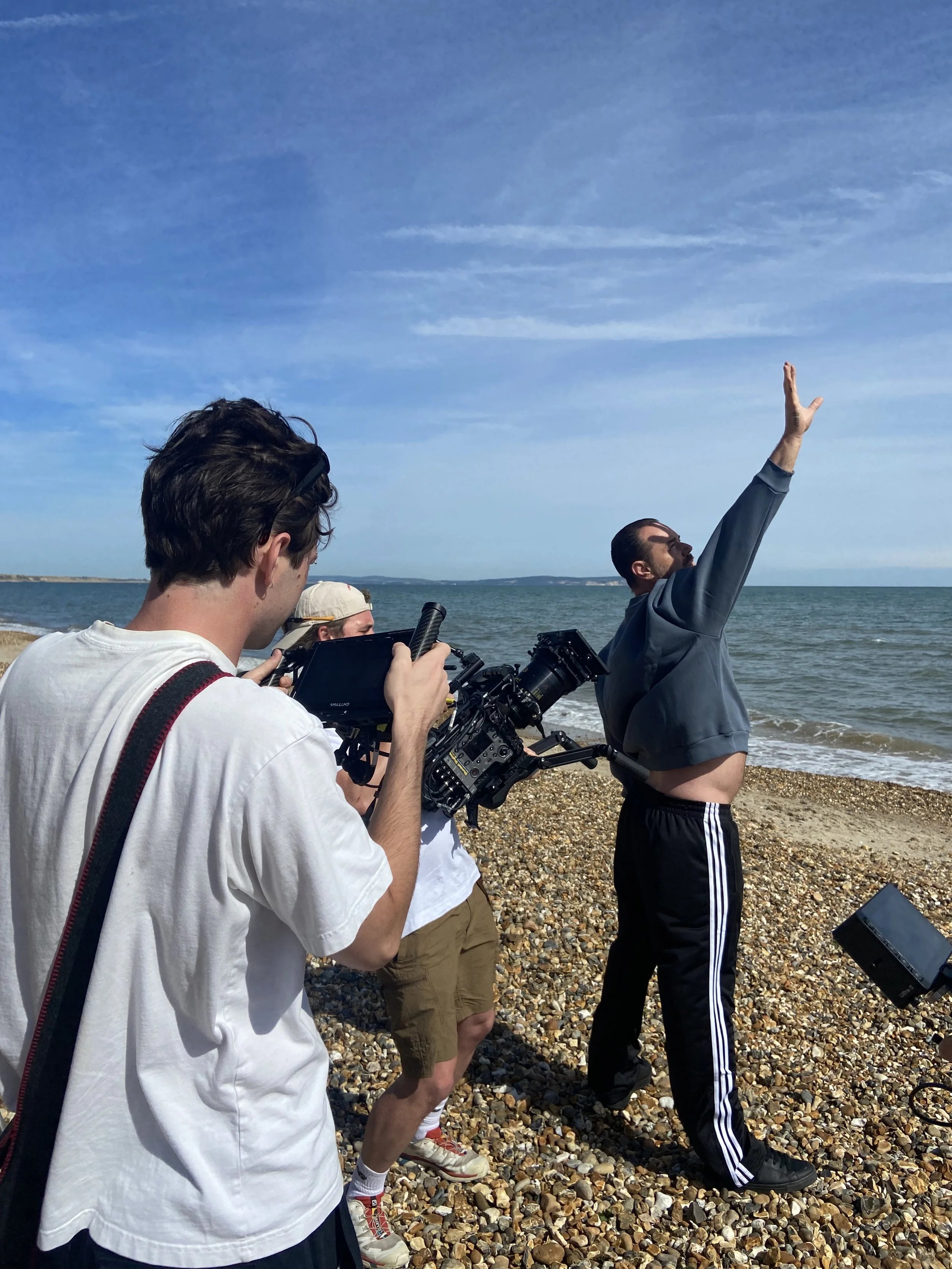 A group of people filming on a beach, with a man in a gray hoodie and black Adidas pants striking a dramatic pose by the water.