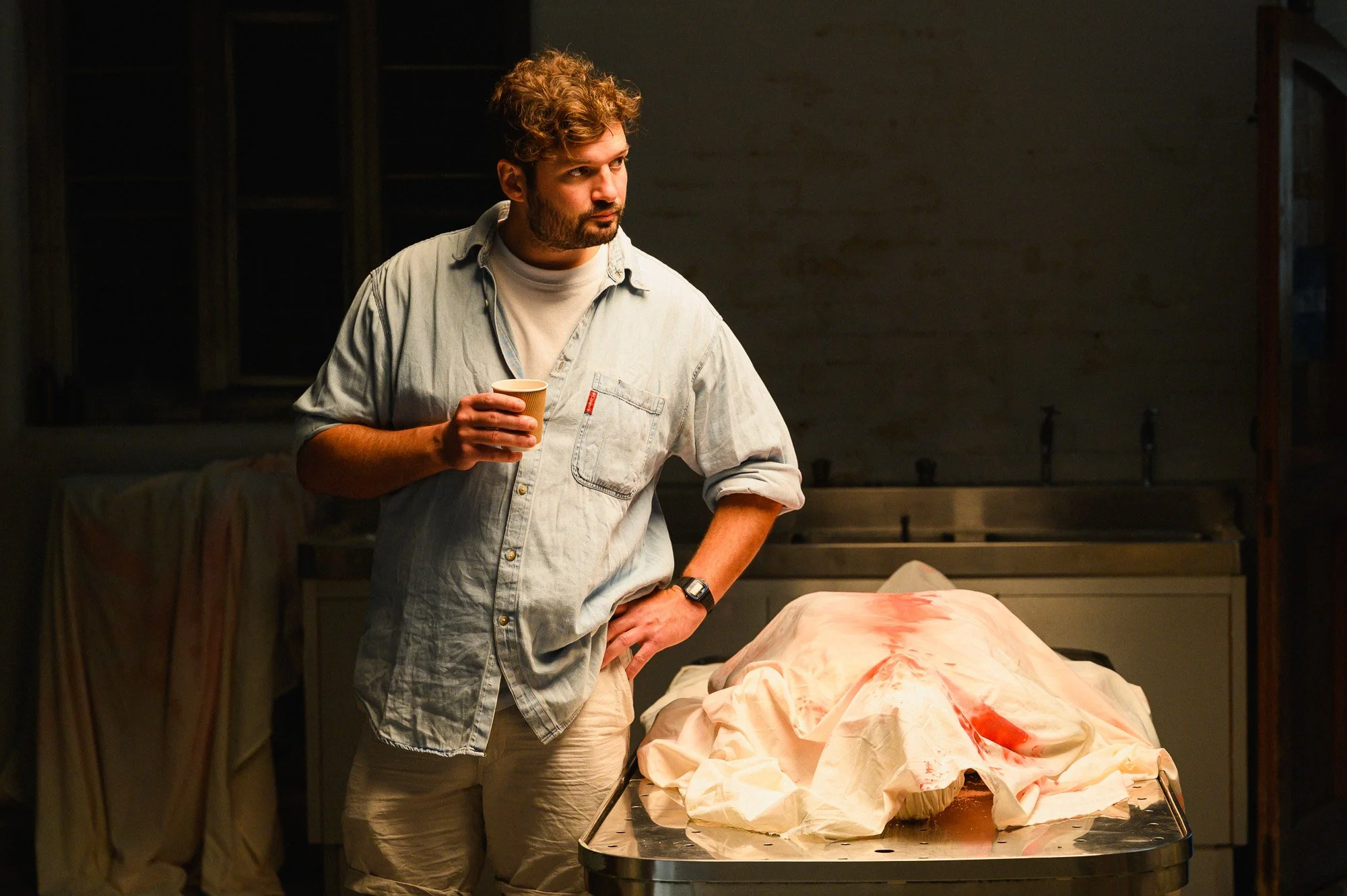 A man with curly hair and beard holding a cup, standing in a dimly lit room with a covered body on a table in front of him.