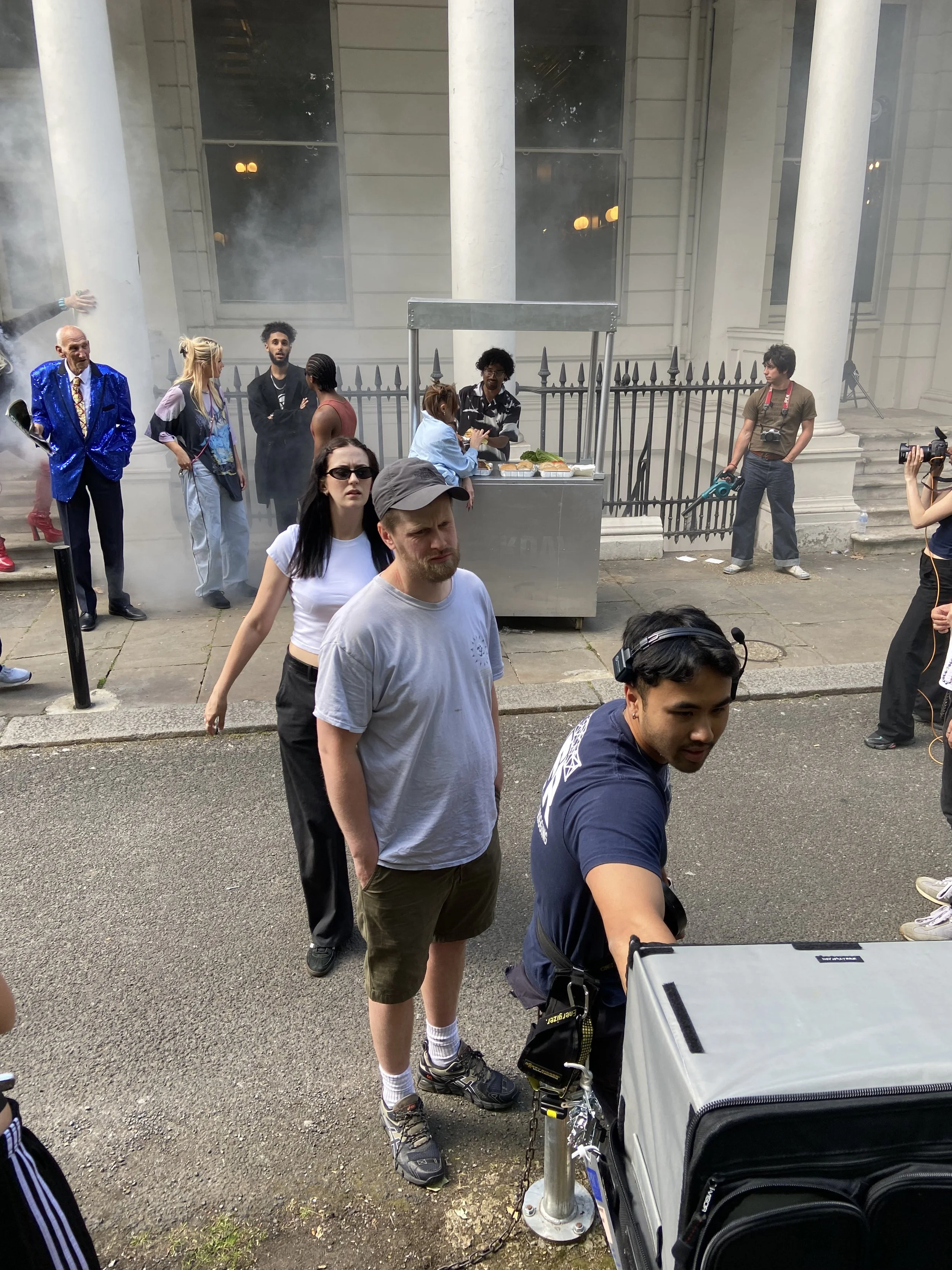 People standing in line outside a building, with crew members preparing for filming or photography, and grill cart in the background.