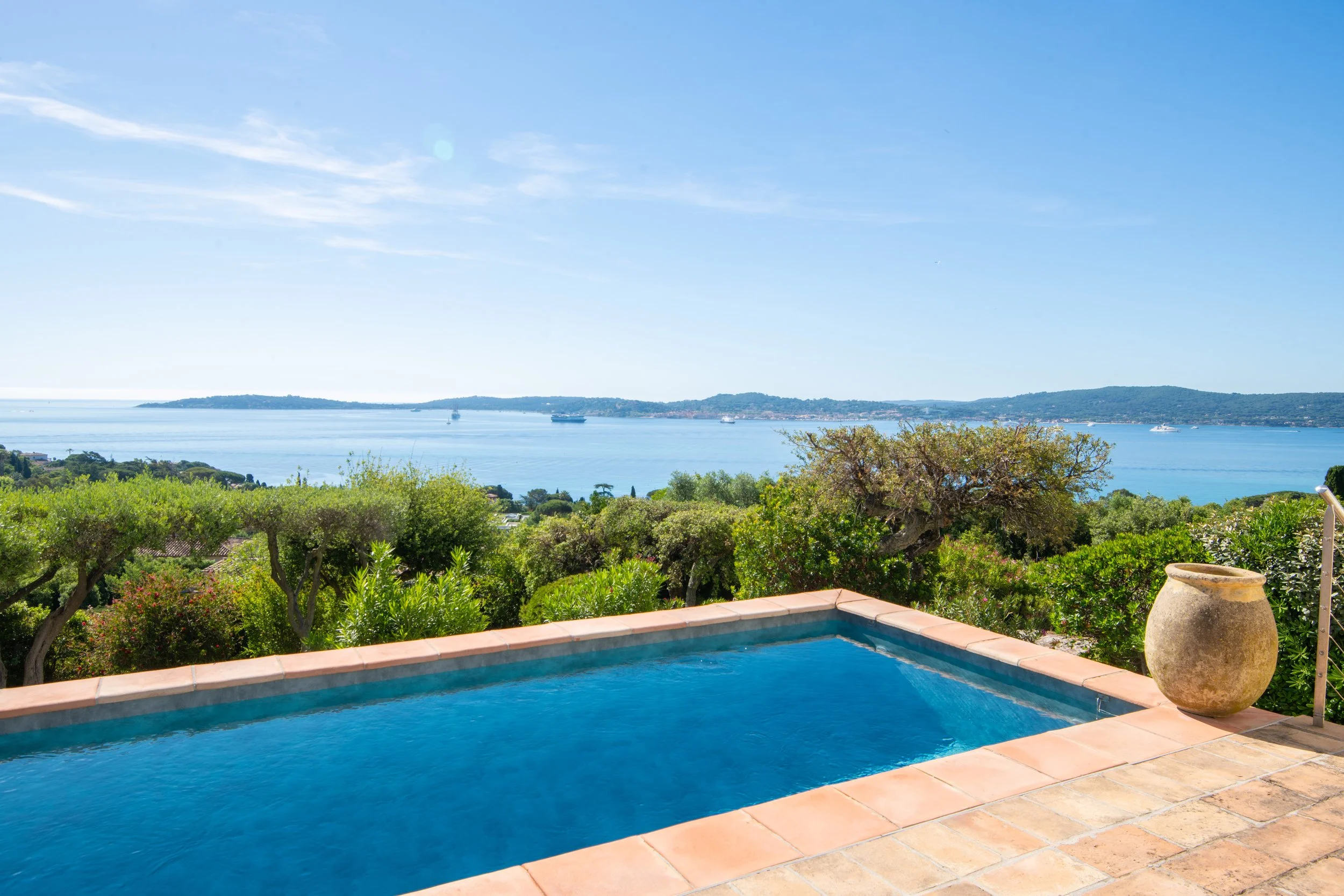 Vue panoramique d'une piscine avec en arrière-plan une mer bleue étincelante, des collines verdoyantes et des voiliers sur l'eau par temps clair et ensoleillé.