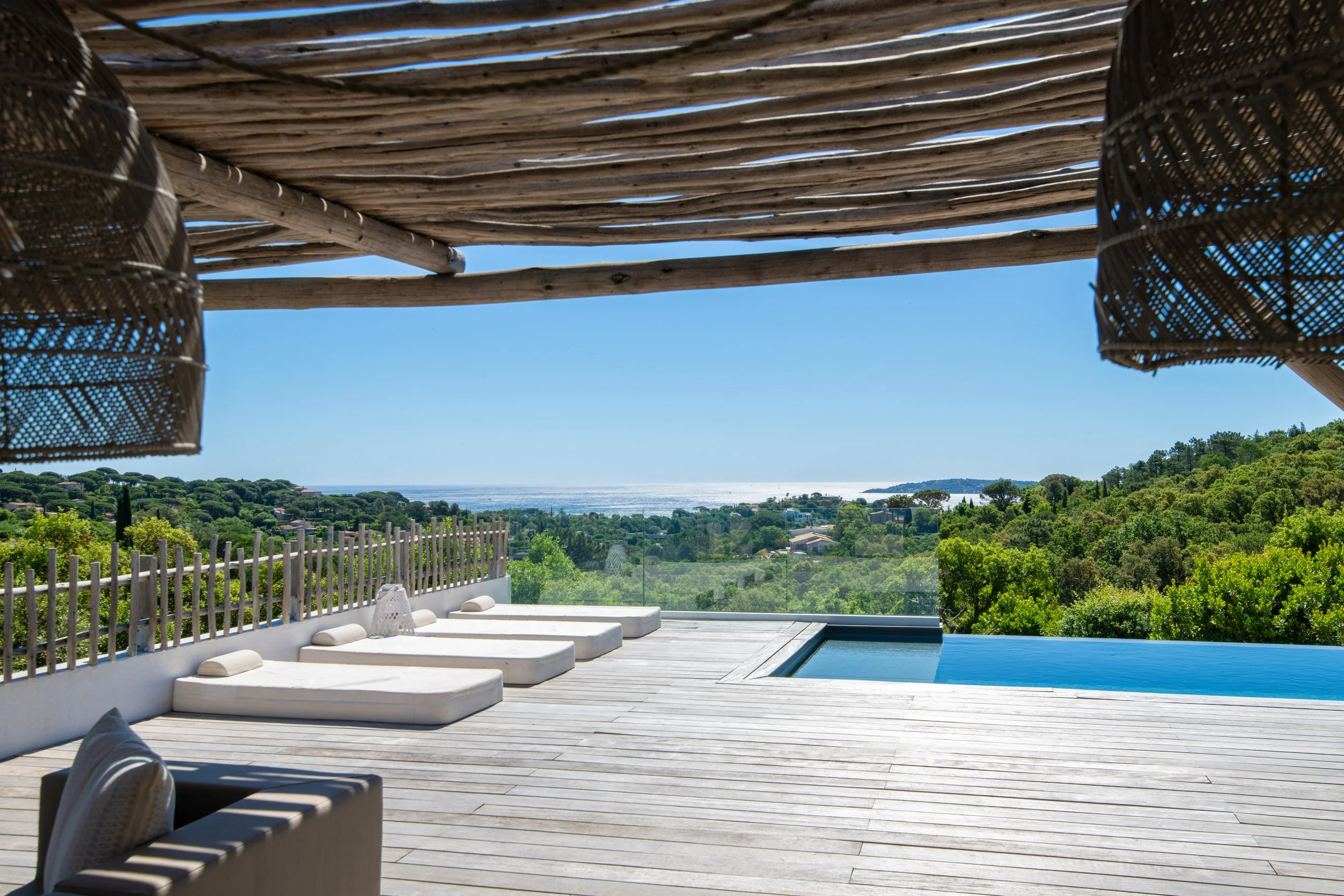 Vue d'une terrasse en bois avec une piscine, entourée de collines verdoyantes et d'une vue sur l'océan à l'horizon sous un ciel bleu.