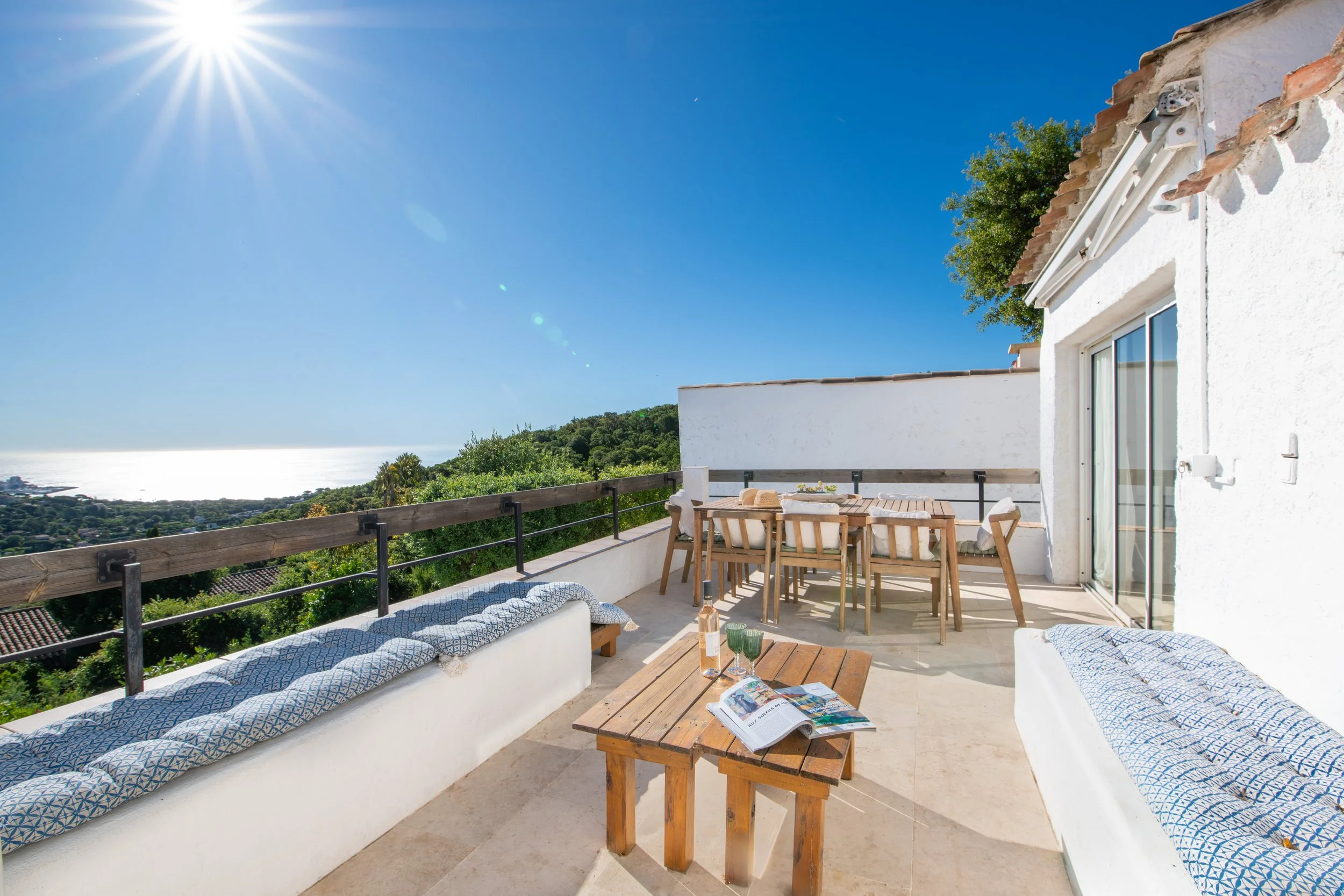 Terrasse ensoleillée avec vue sur la mer, meubles en bois, table et sièges, avec une décoration simple et végétation environnante.