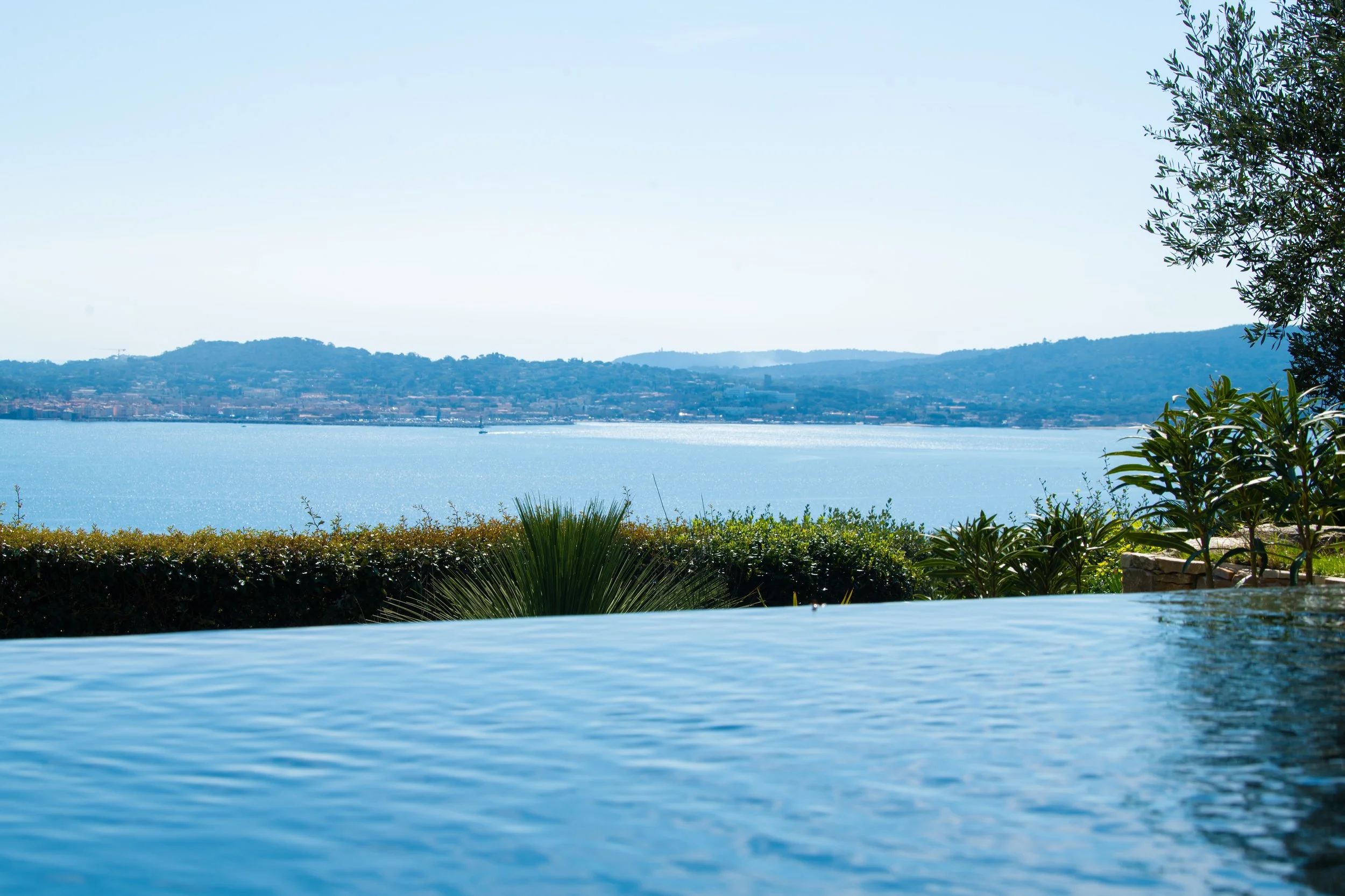 Vue sur la mer avec la plage et des collines au loin depuis une piscine avec un paysage de jardin soigné.