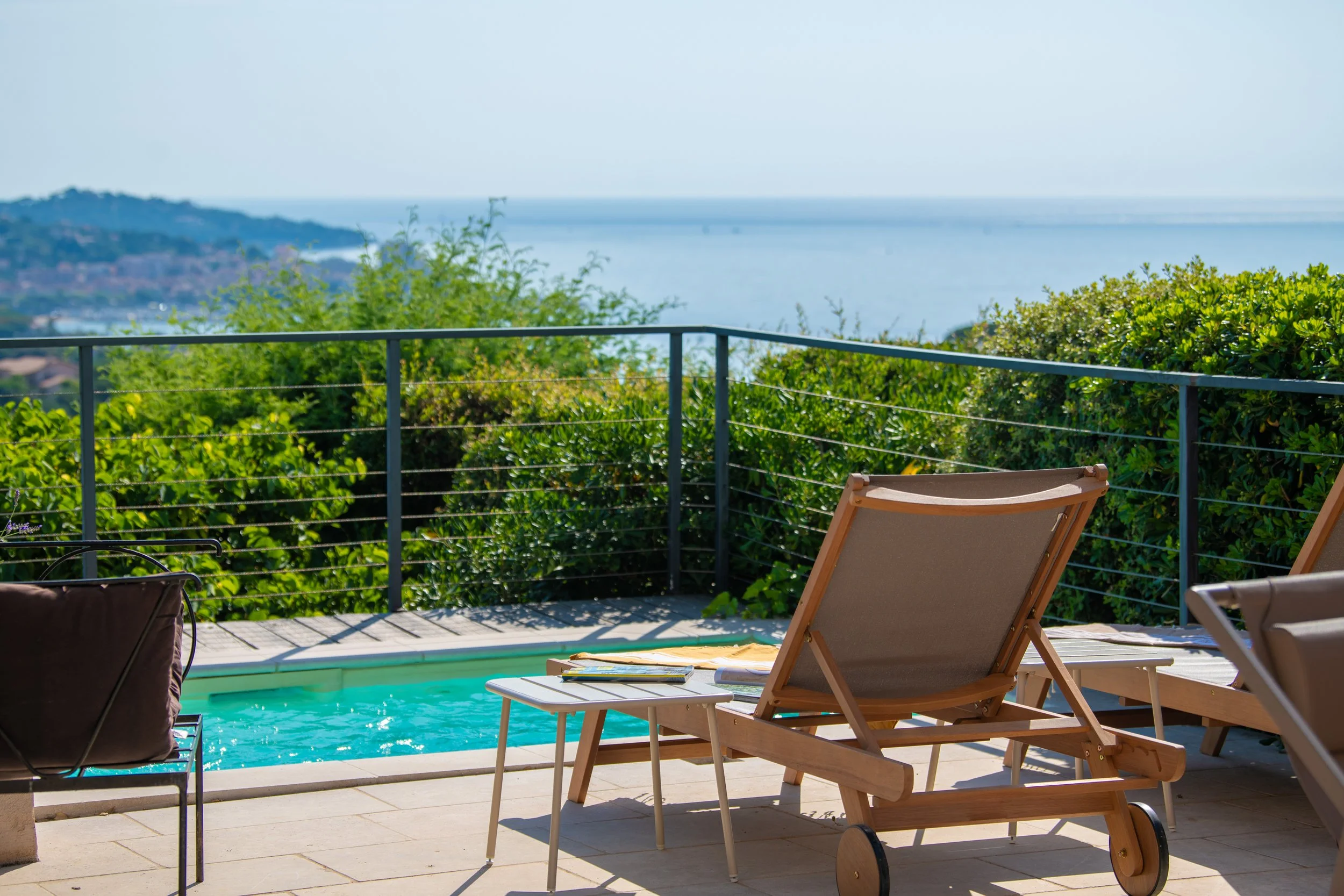 Terrasse avec piscine surplombant la mer et la côte, transats, table avec bouteille de vin et verre, ciel bleu.