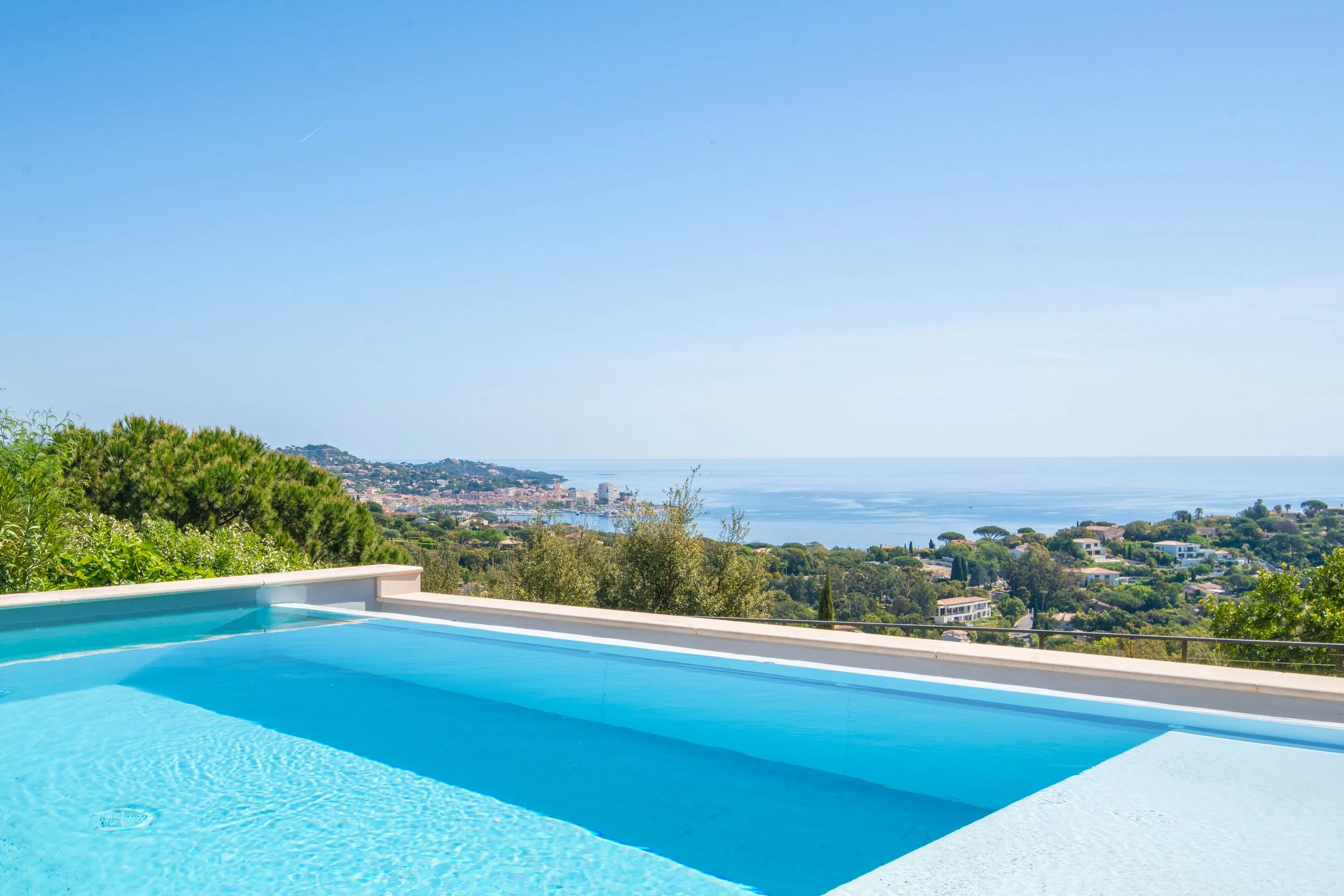 Vue d'une villa moderne avec piscine donnant sur la mer et les collines environnantes, ciel bleu dégagé.