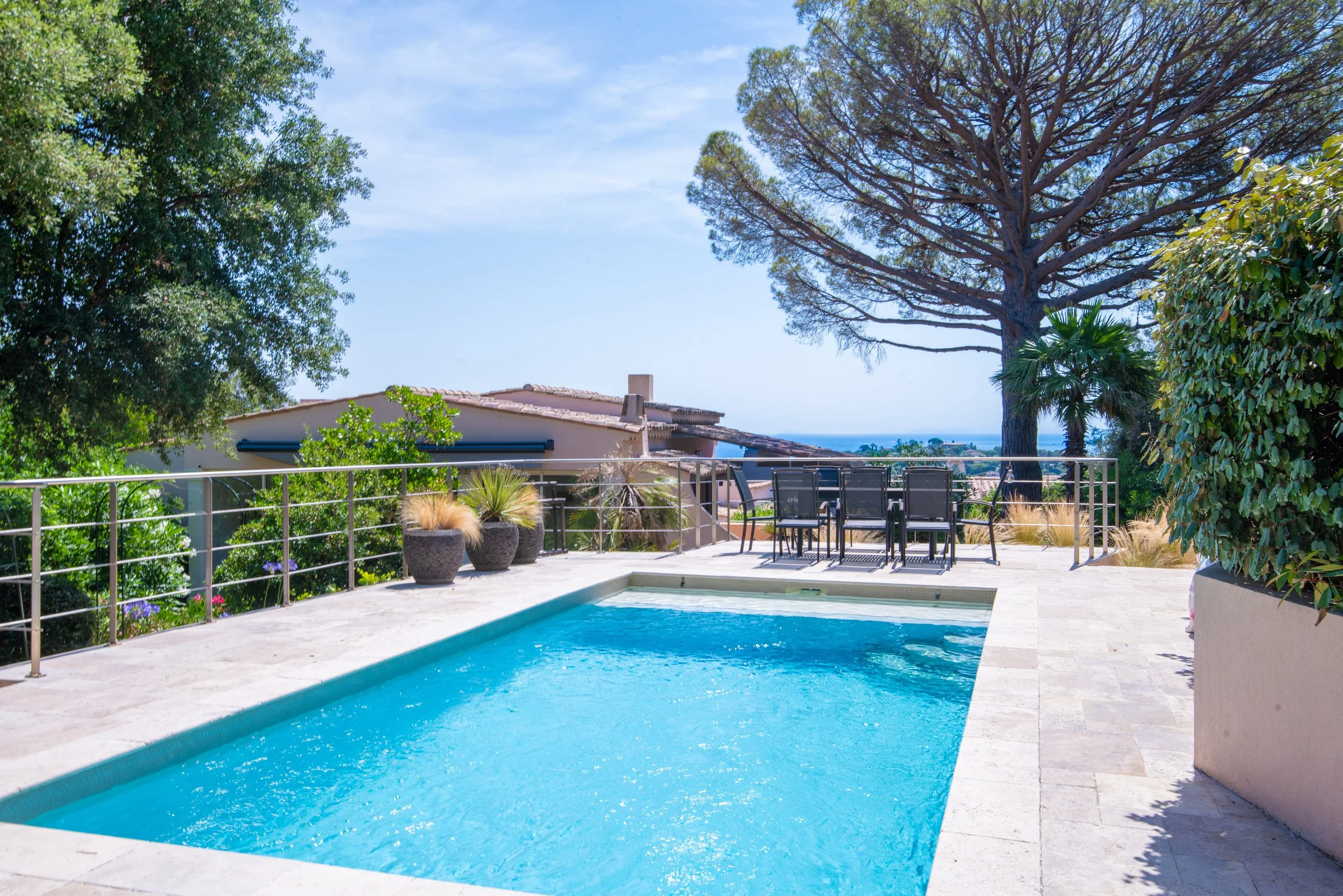 Piscine extérieure entourée de carrelage, avec arbres et mobilier de jardin en arrière-plan, sous un ciel bleu.