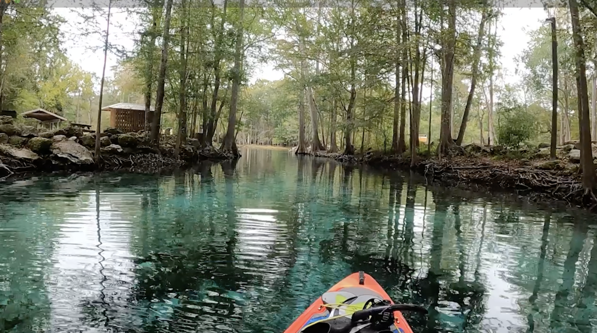 View from a kayak paddling through a calm, clear waterway surrounded by dense trees and rocky banks with small wooden structures on the shore.