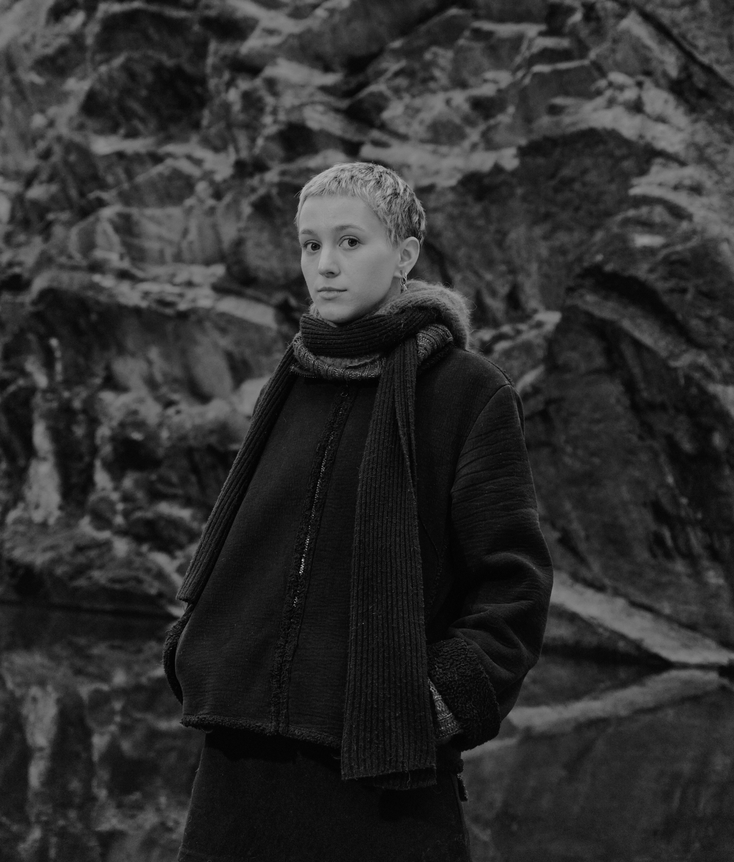 Svart-hvít portrett af ungri konu með stutt hár stendur úti fyrir framan grýttan bakgrunn, klædd í dökkan jakka og trefil.