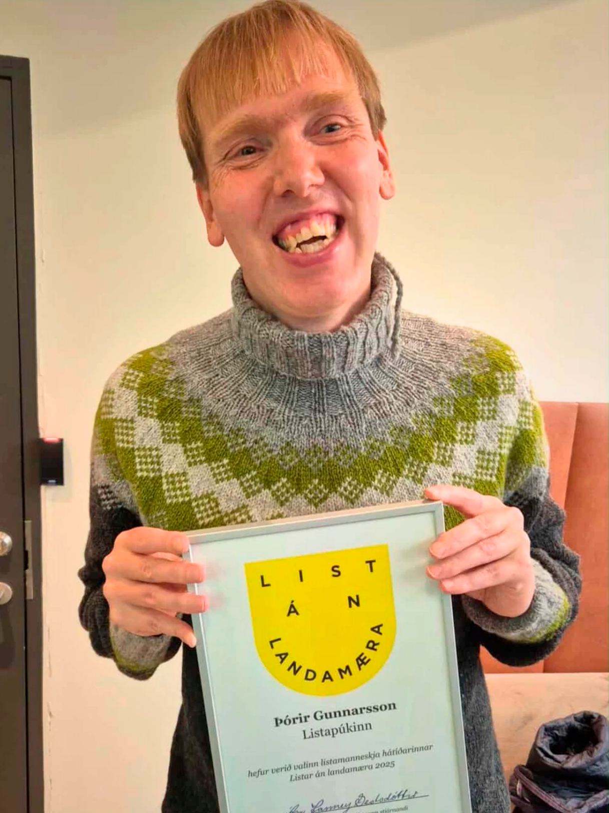 Listapúkinn brosandi í fallegri lopapeysu heldur á viðurkenningarskjali fyrir að vera listamanneskja List án landamæra 2025