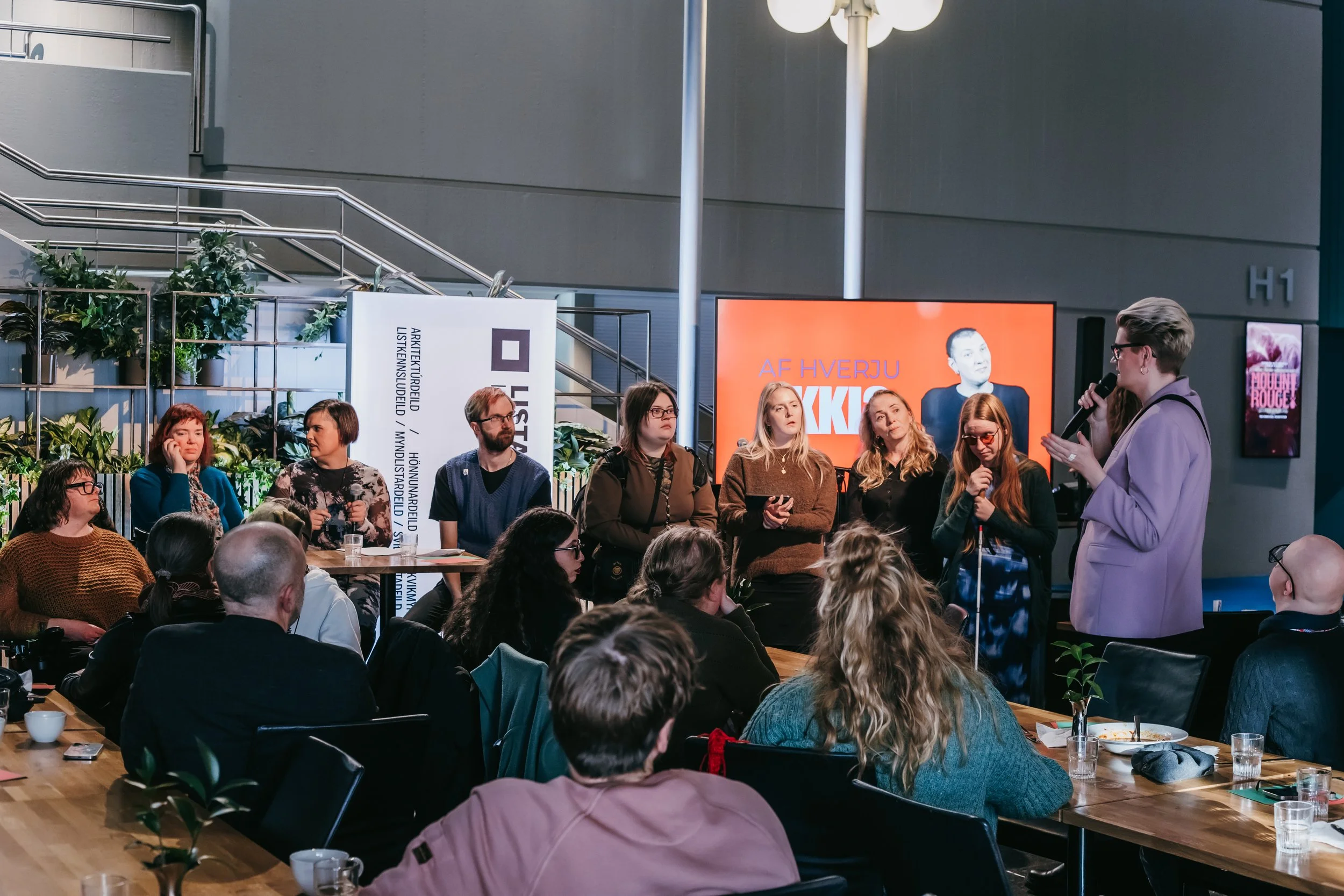 Fólk situr í röð á sviðinu, stór skjár í bakgrunninn. Spjallstjórinn talar í hljóðnema, fólk í salnum situr og horfir á.