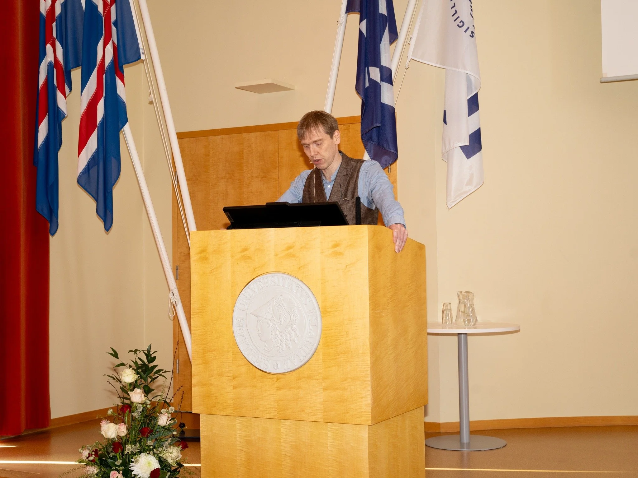 Maður að lesa úr pontu, fánar til hliðar við hann og blómvöndur.