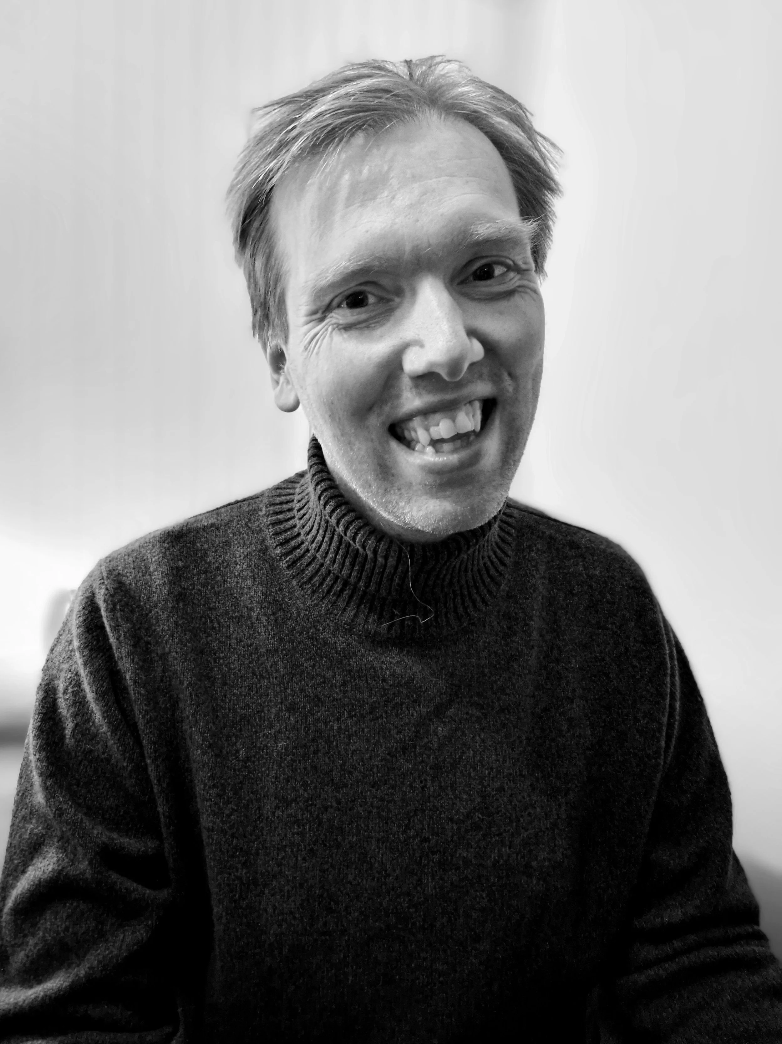 Black and white photo of a smiling man with short hair, wearing a dark sweater, against a plain light background.