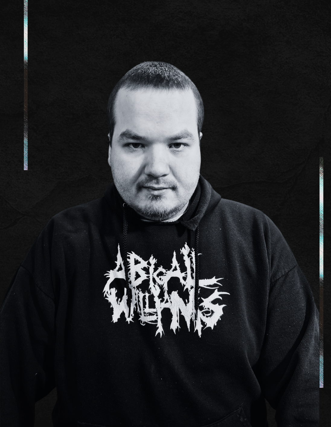 A young man with short dark hair, light facial hair, and a serious expression posing against a dark background. He is wearing a black hoodie with a white graphic design on the front.