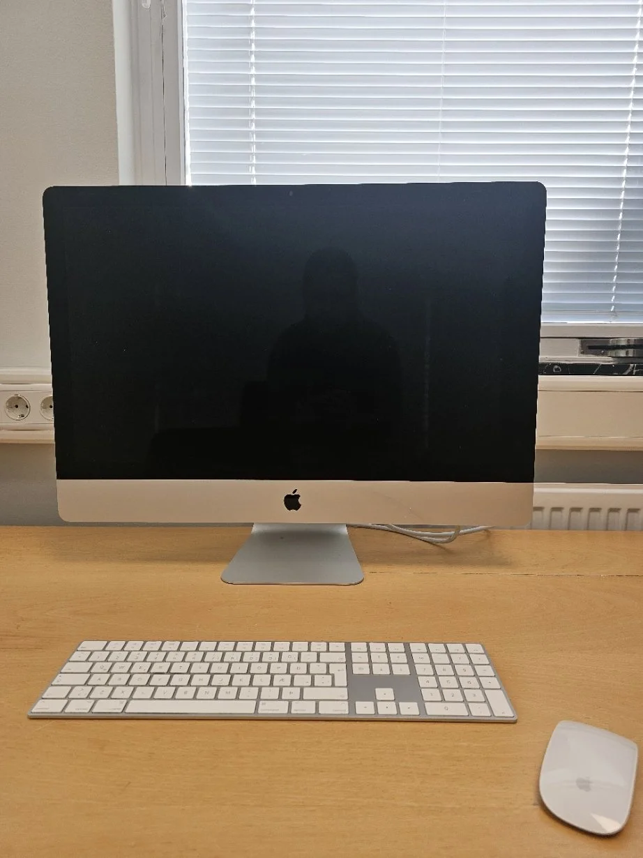 Apple iMac-tölva með lyklaborði og mús á viðarborði. Gluggi með rimlagardínum er í bakgrunni.