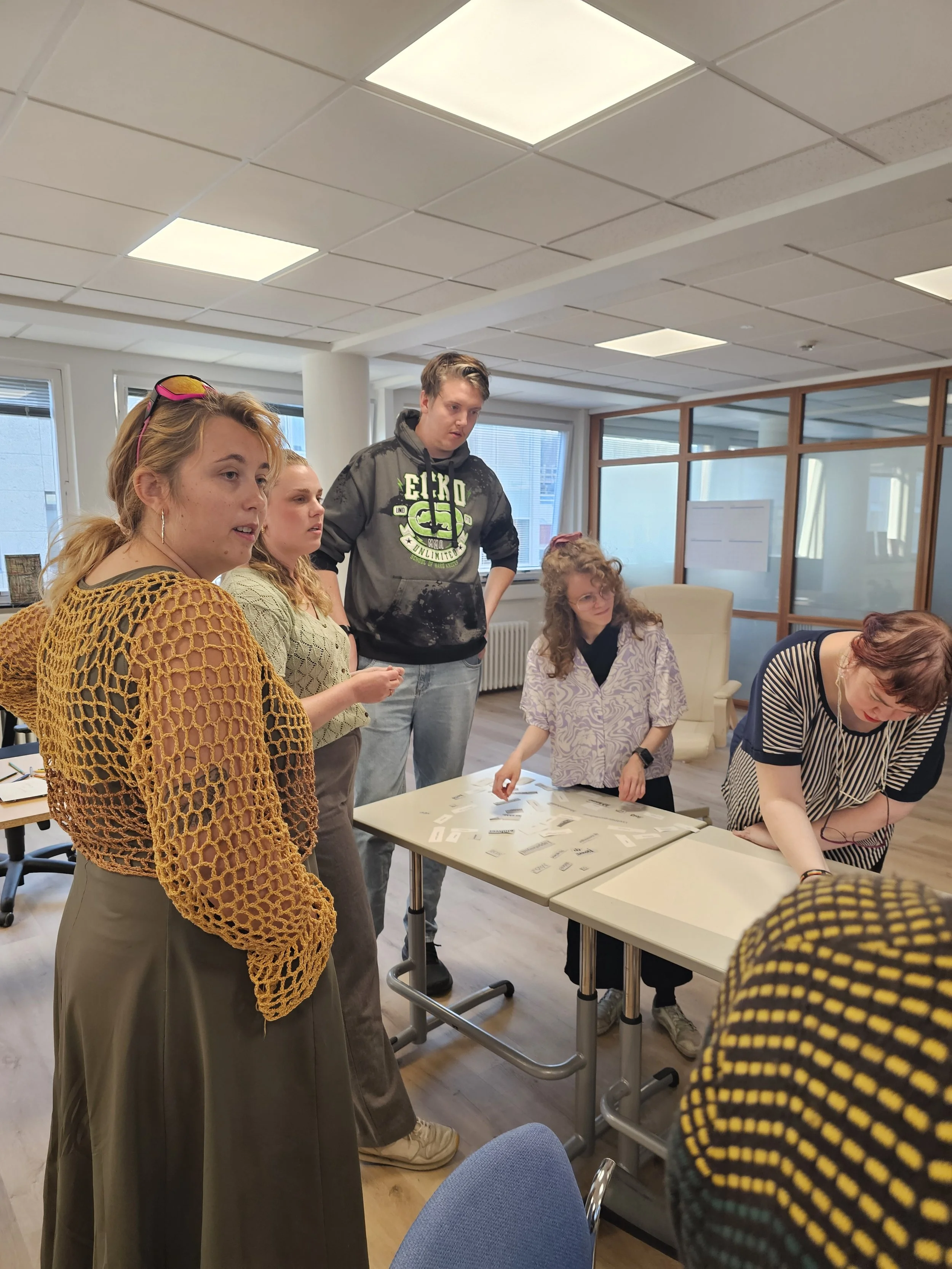 Fimm manna hópur saman við borð sem vinnur með pappír og útklippur. Þau eru stödd í vel upplýstu herbergi með stórum gluggum og hvítum flísum í loftinu.