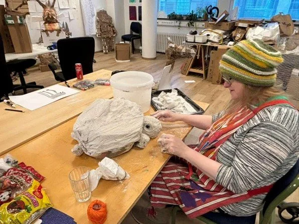 A woman wearing a striped sweater, glasses, and a knit hat working on a sculpture of a turtle on a wooden table filled with art and craft supplies in an art studio.