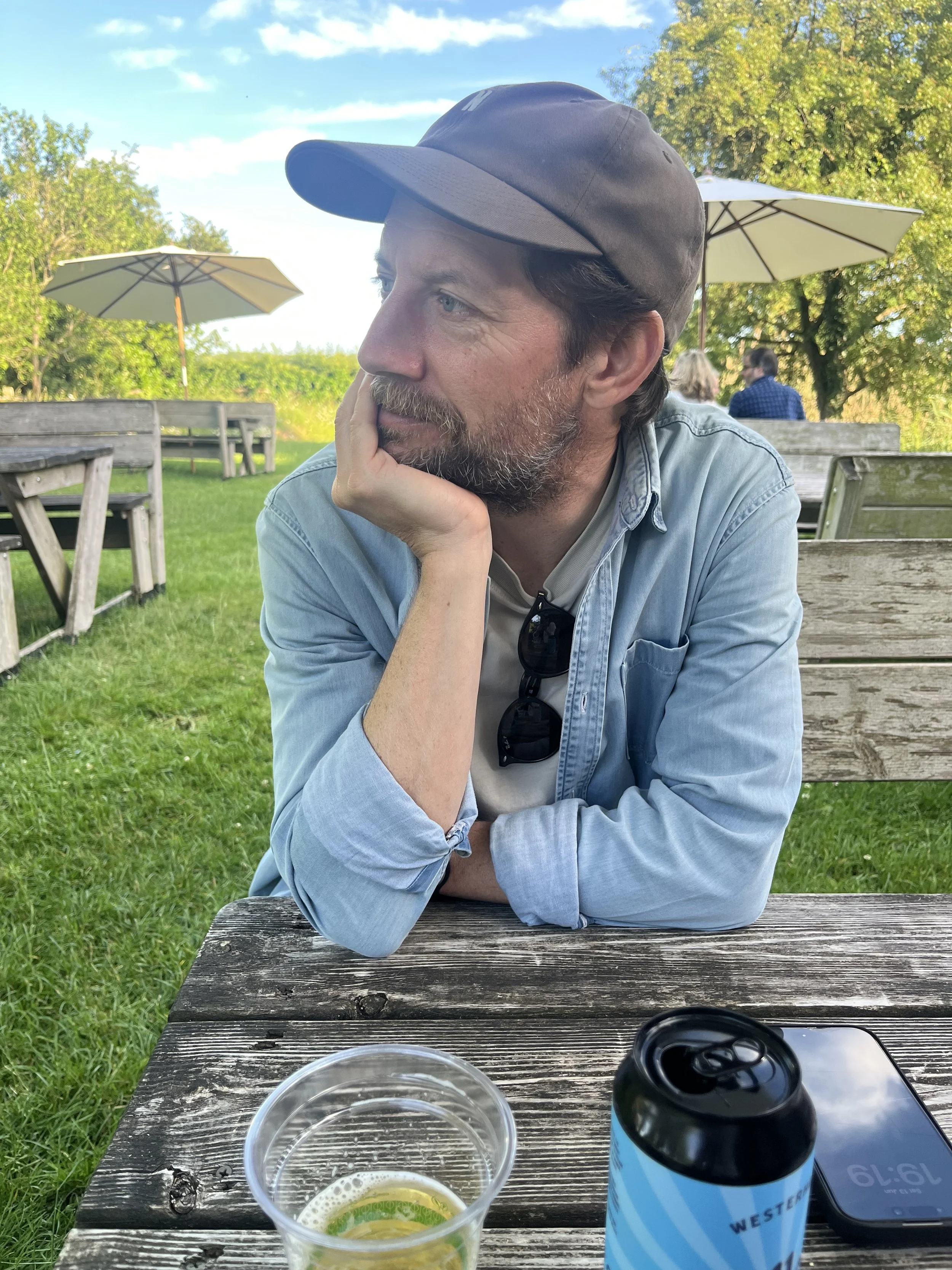A man with light skin, a beard, and blue eyes, wearing a black cap and a light denim shirt, sits at a weathered wooden outdoor table with his head resting on his hand, in a park with trees, umbrellas, and people in the background. On the table are a plastic cup with a lime slice and a dark-colored can.