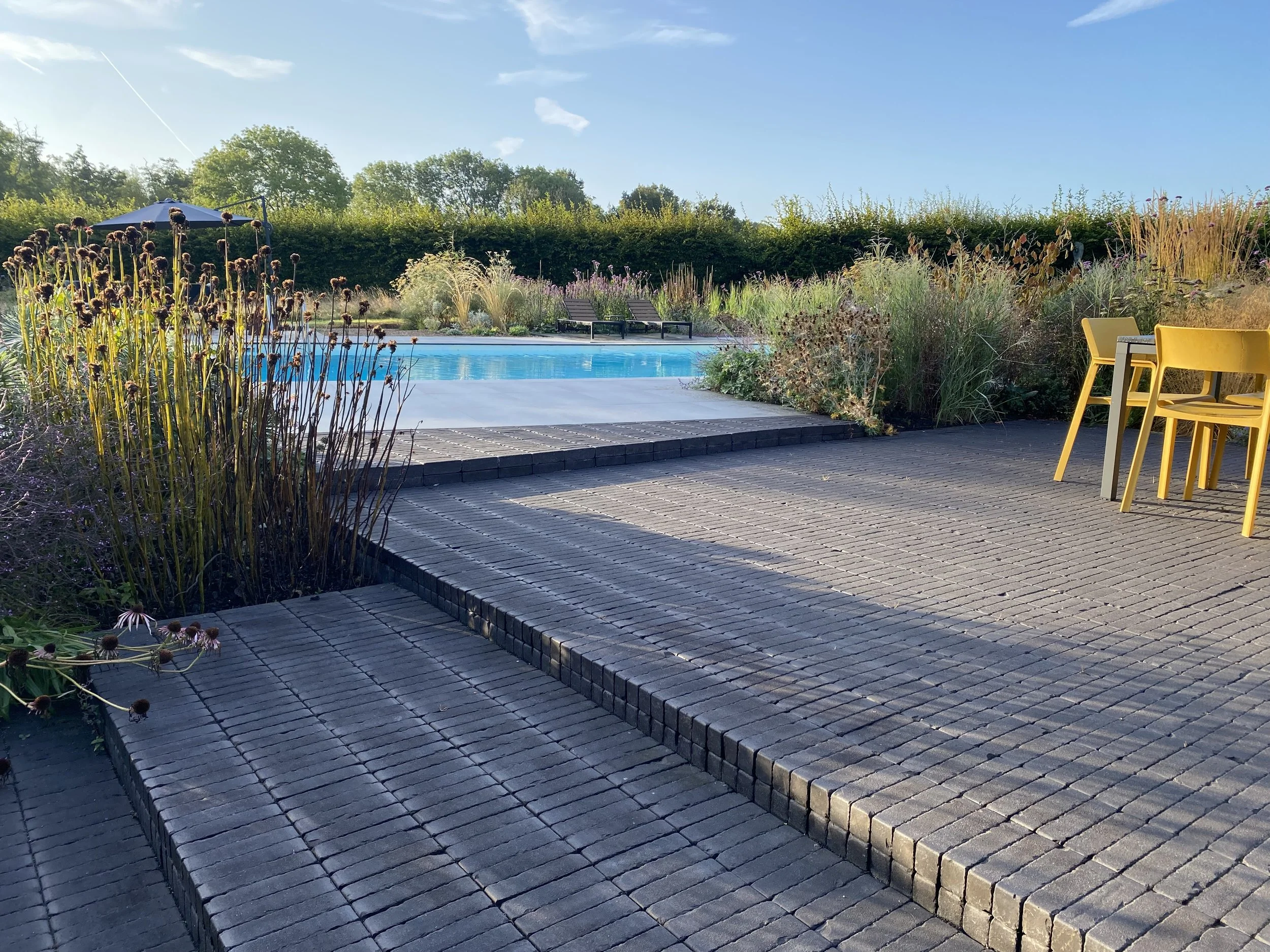 An outdoor scene featuring a swimming pool surrounded by a wooden deck, yellow outdoor chairs, lush greenery, and a blue sky with some clouds.