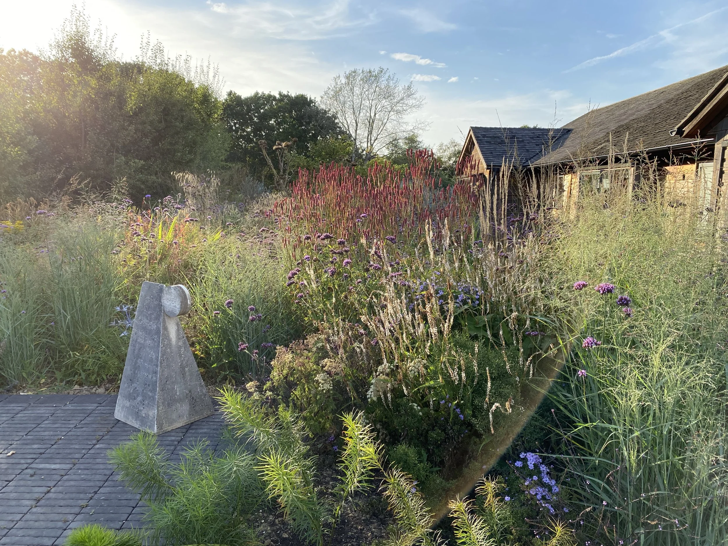 A garden scene with various colorful flowers and tall grasses, a concrete sculpture in the foreground, and a house with a shingled roof in the background. The sky is partly cloudy with sunlight illuminating the scene.