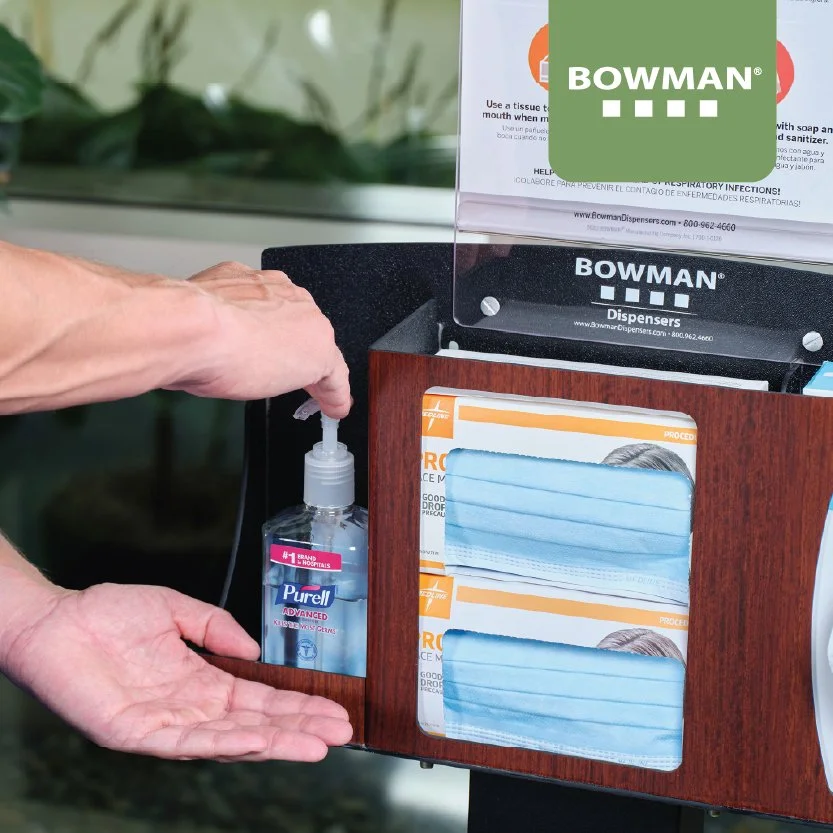 Person's hand reaching for a bottle of Purell hand sanitizer placed inside a wooden dispenser station, which also contains disposable face masks and informational pamphlets.