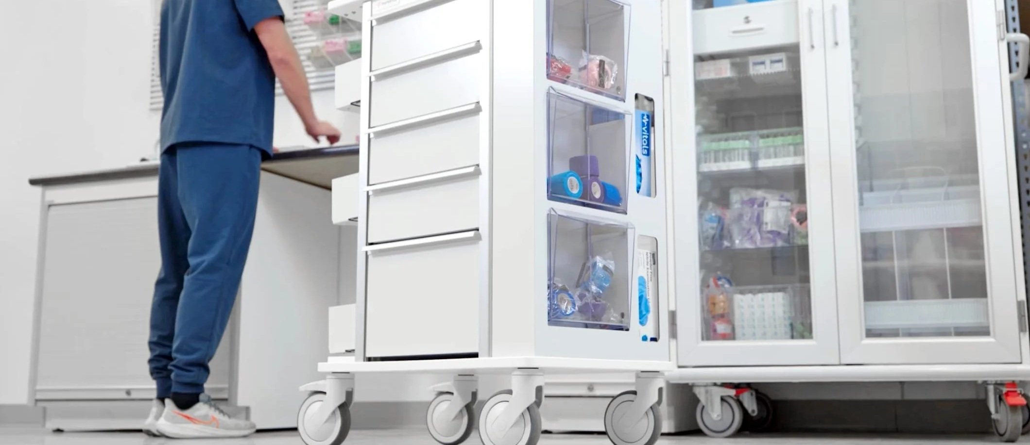 Person in blue scrubs pushing a hospital or pharmacy cart with transparent refrigerated compartments, stocked with medical supplies in a clinical setting.