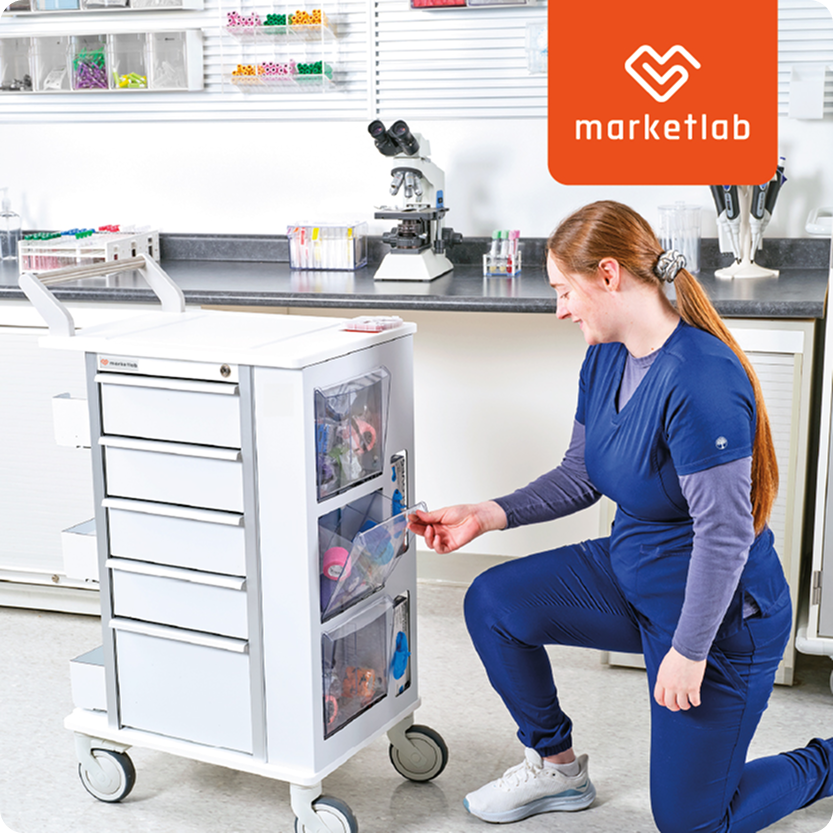 A woman in blue medical scrubs kneeling next to a white medical cart in a laboratory, with a microscope and shelves with supplies in the background.