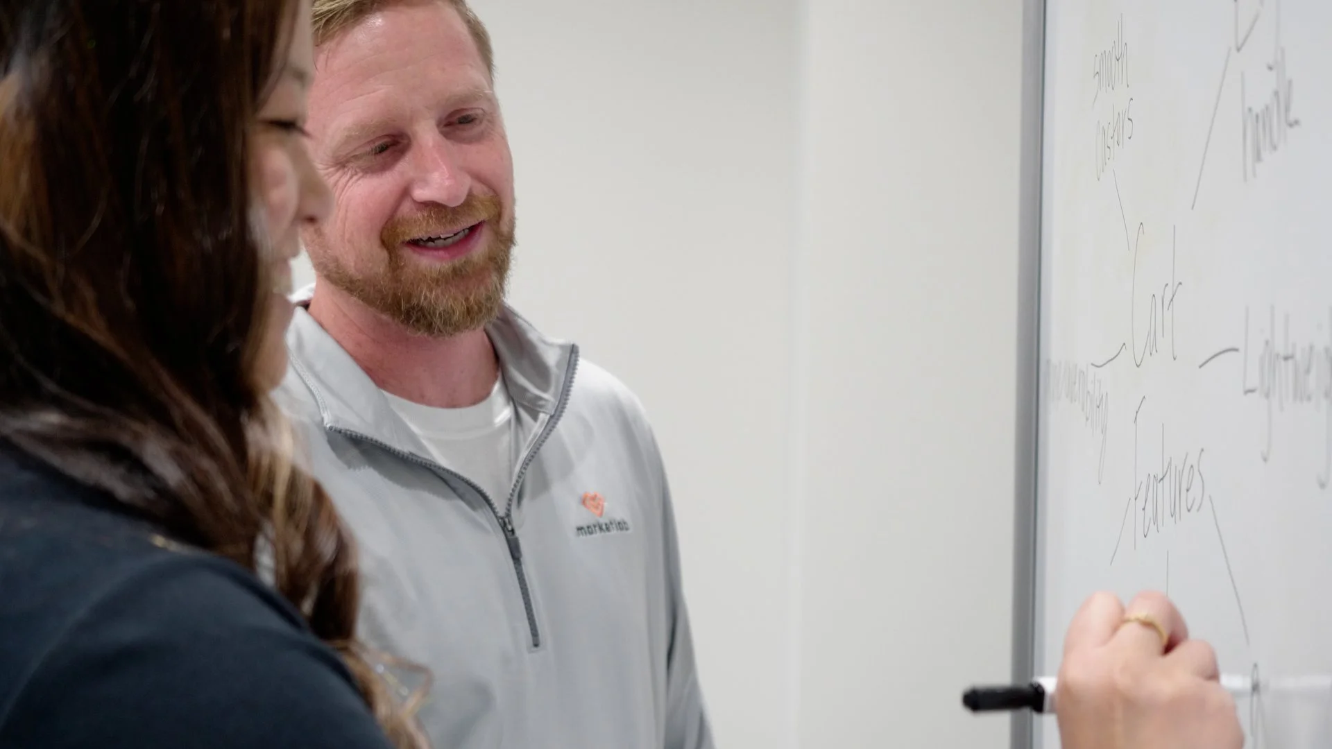 Two people, a woman with long brown hair and a man with a beard, are smiling and writing on a whiteboard in a room with white walls.