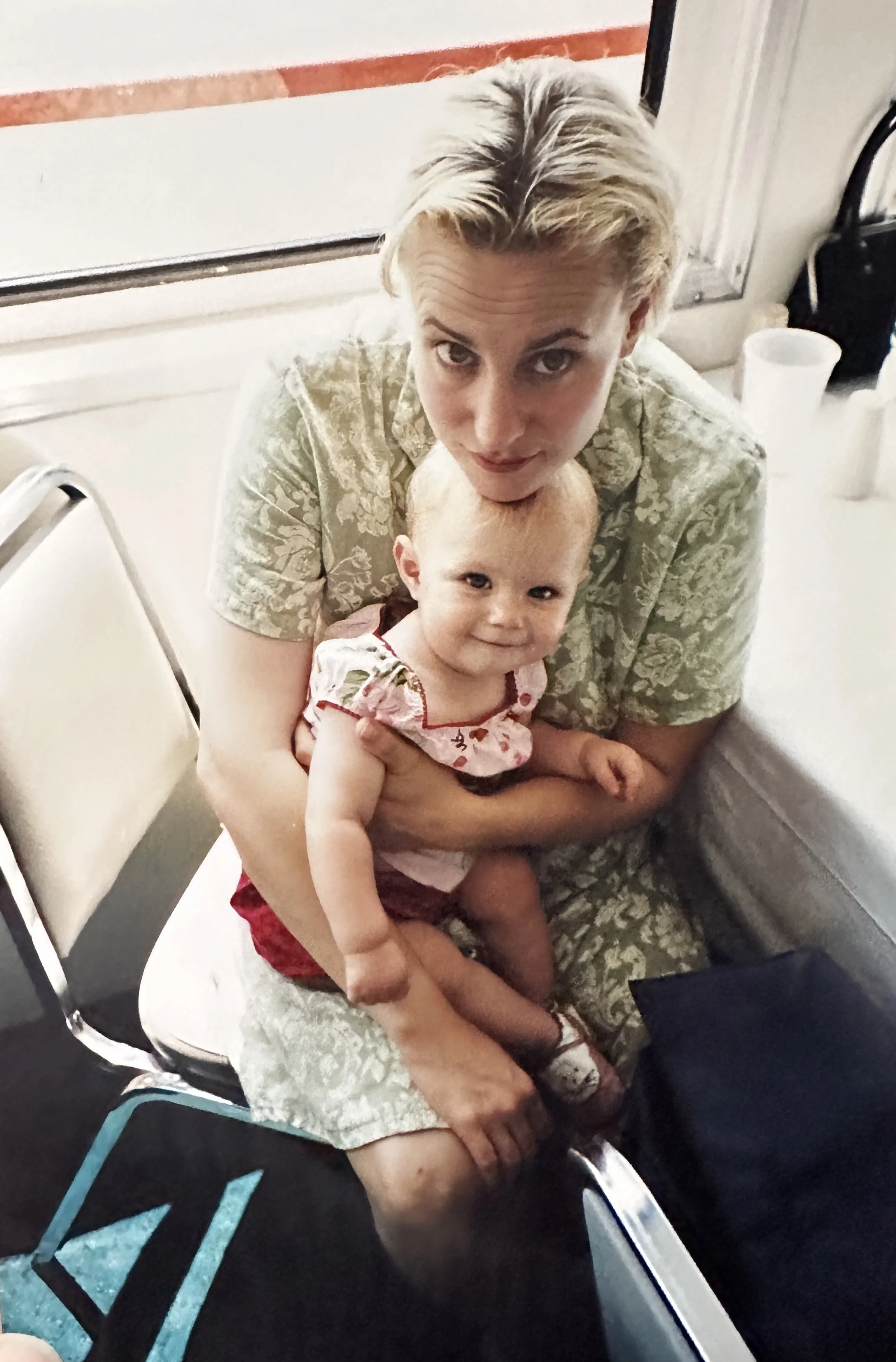 A woman sitting on a chair holding a smiling young baby on her lap. The woman has short blonde hair and is wearing a patterned dress, while the girl is dressed in a pink and red outfit. They are in an indoor setting near a table and chairs.