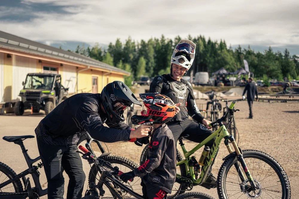 Två personer med mountainbikes, en av dem håller på att få på sig hjälm, den andra bär hjälm på huvudet och ler, utomhus på en bike park.
