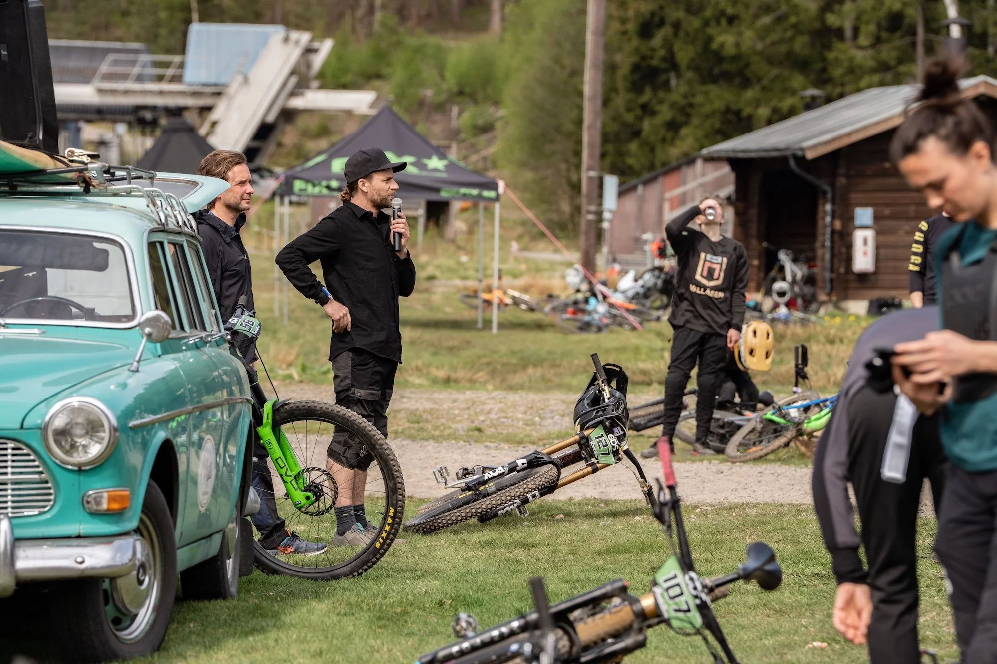 Människor samlas utomhus med mountaincyklar, en man håller en mikrofon och talar, andra förbereder eller vilar med cyklar, en gammal blå bil är parkerad bredvid, en svart tält står i bakgrunden, och det finns ett pittoreskt, träbyggt hus med skog run