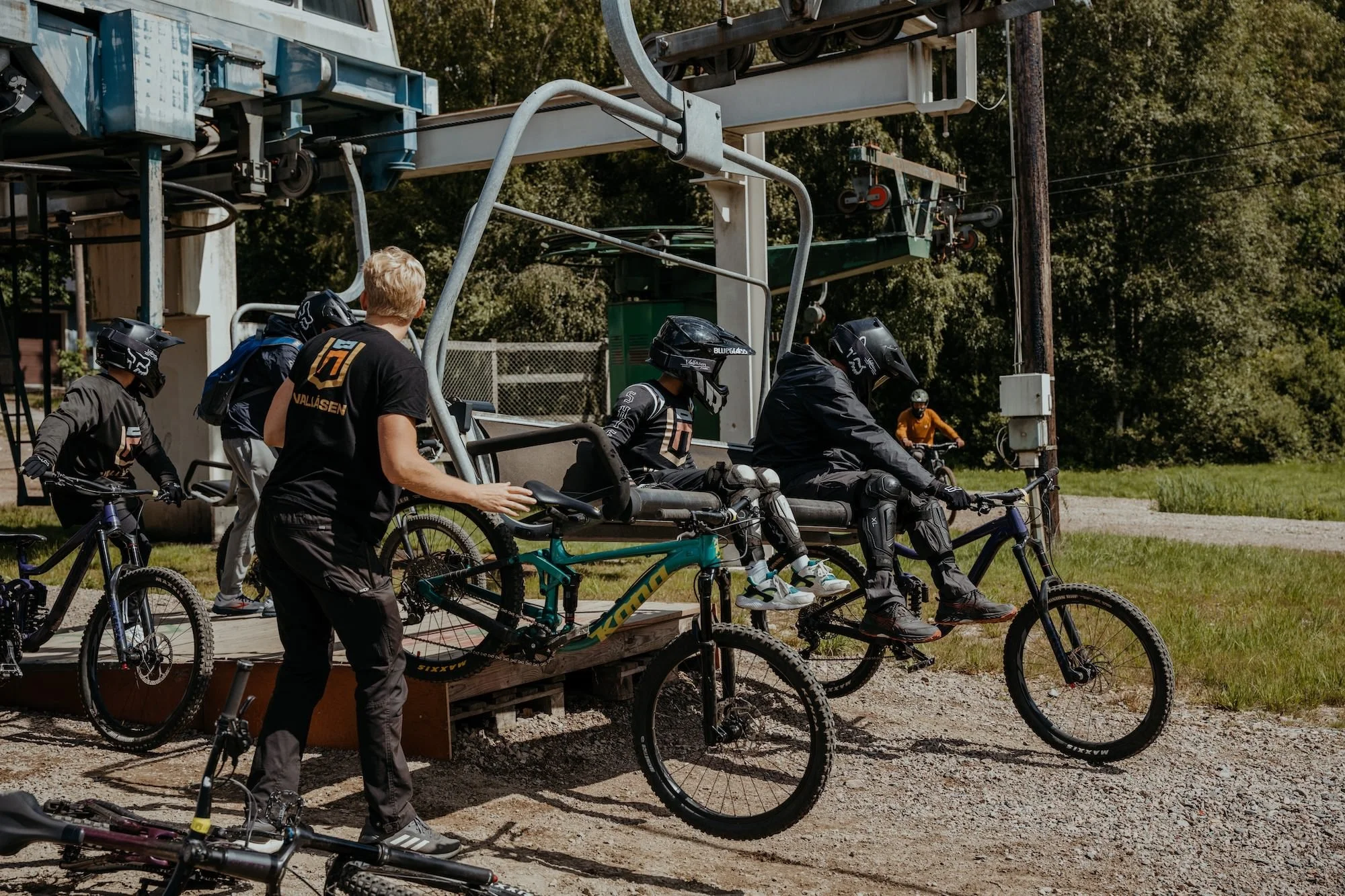 Grupp mountainbikers med hjälmar och skydd förbereder sig för att cykla, några står med cyklar på marken, medan andra sitter på en lift som är del av en cykelpark.