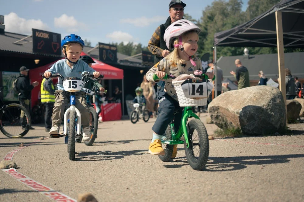 Barn som cyklar på en bana under en cykeltävling med publik och tält i bakgrunden.